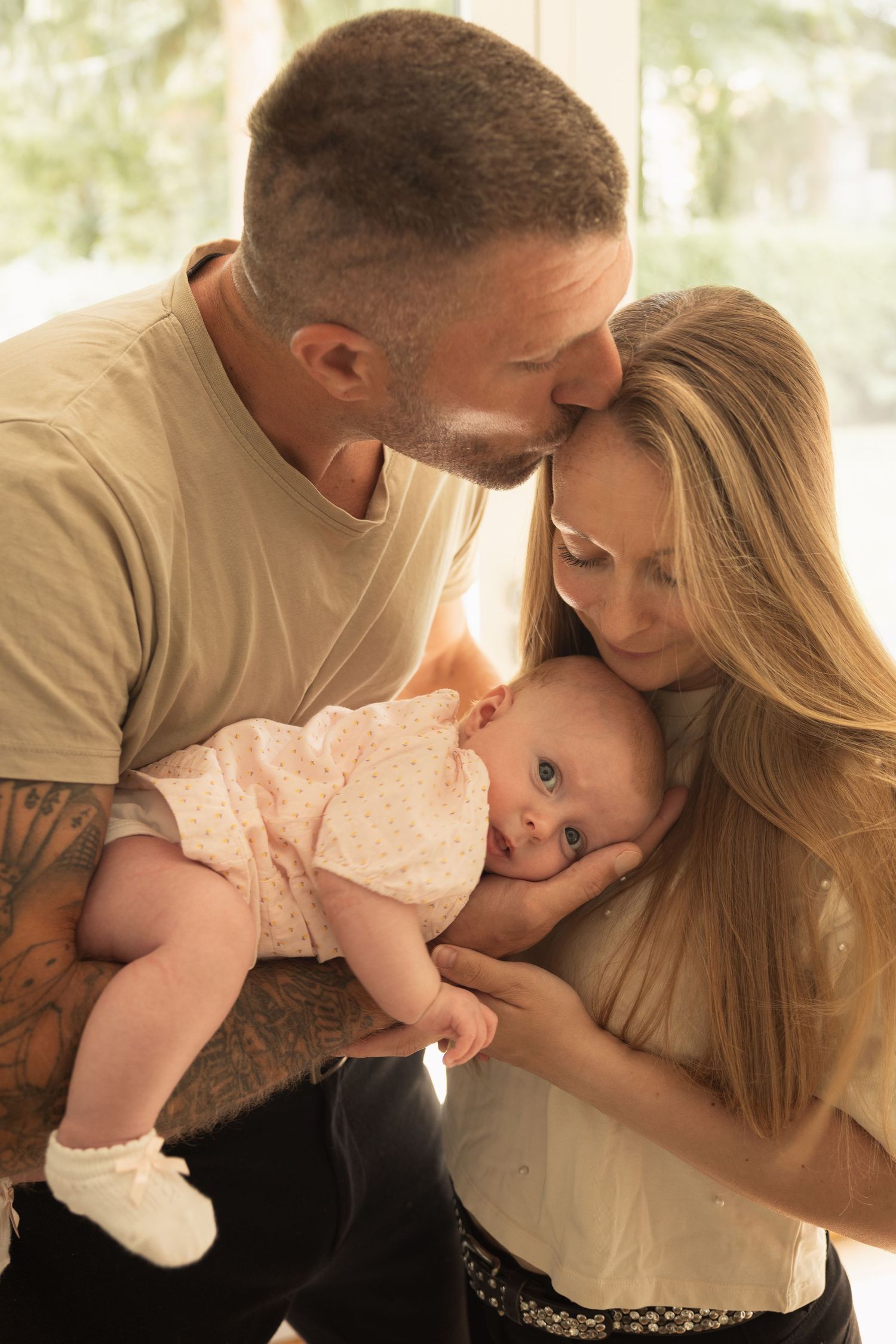 A tattooed father kisses mother's forehead as they both cradle their newborn baby in a warm, sunlit room.