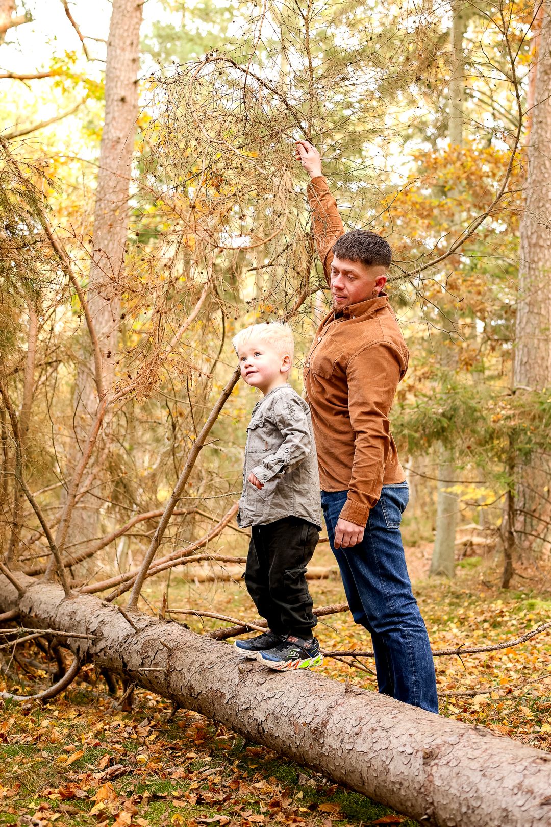 Father helping young blonde boy balance on fallen log in autumn forest.