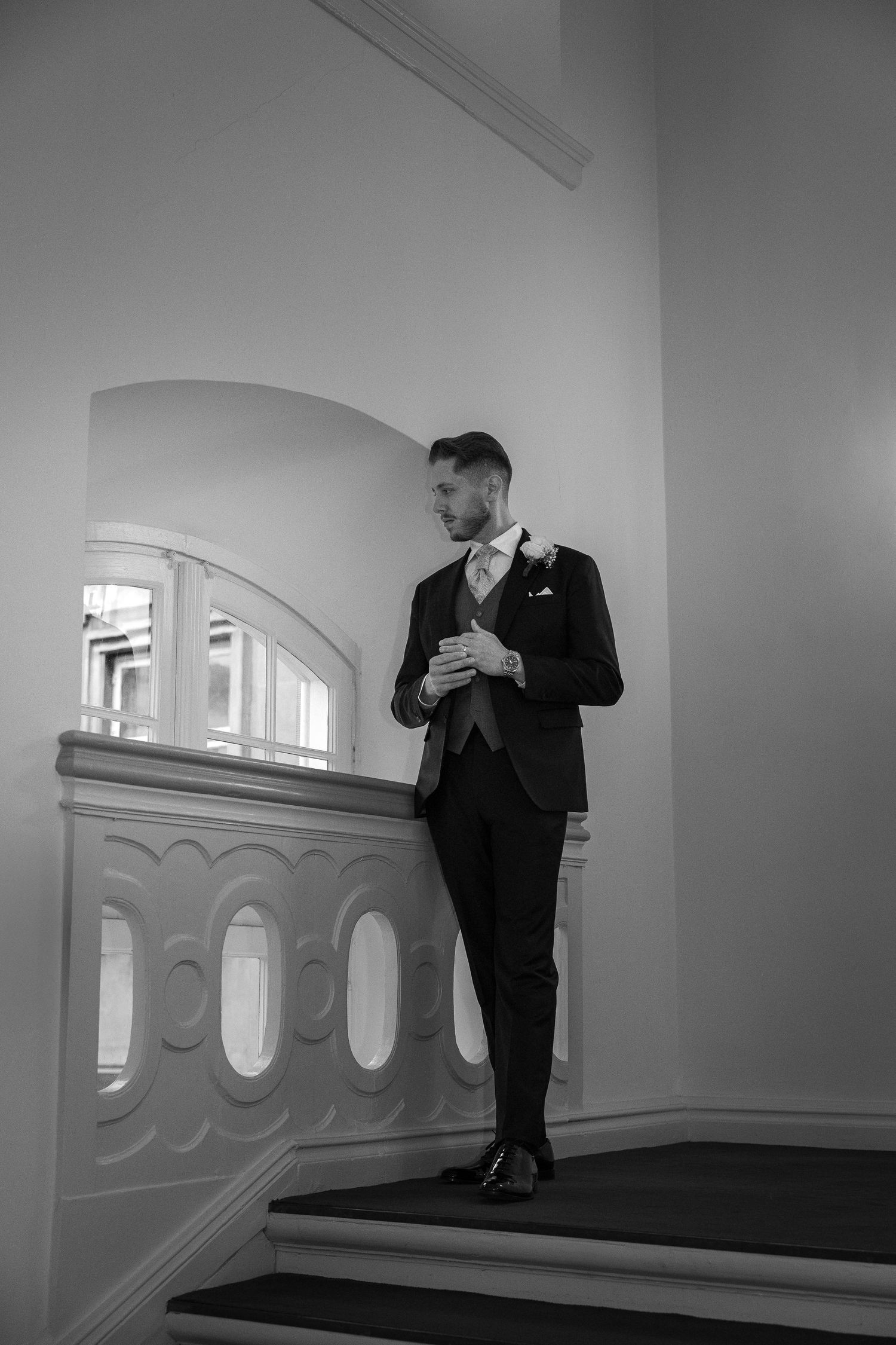 Elegant groom in formal suit stands confidently on ornate staircase in classic black and white photo.