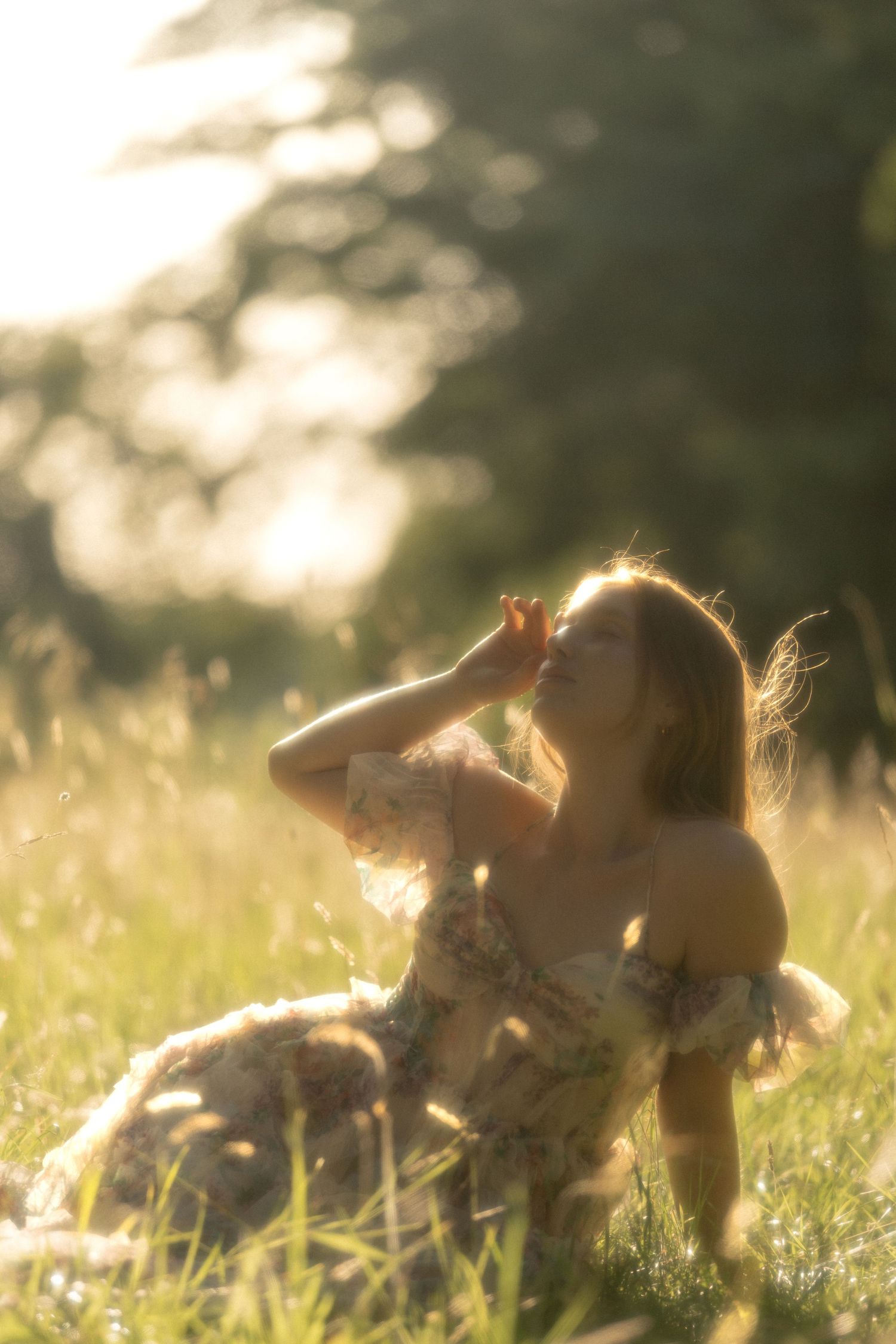 Woman in floral dress sitting in sunlit meadow, golden hour light filtering through trees, hand raised to forehead.