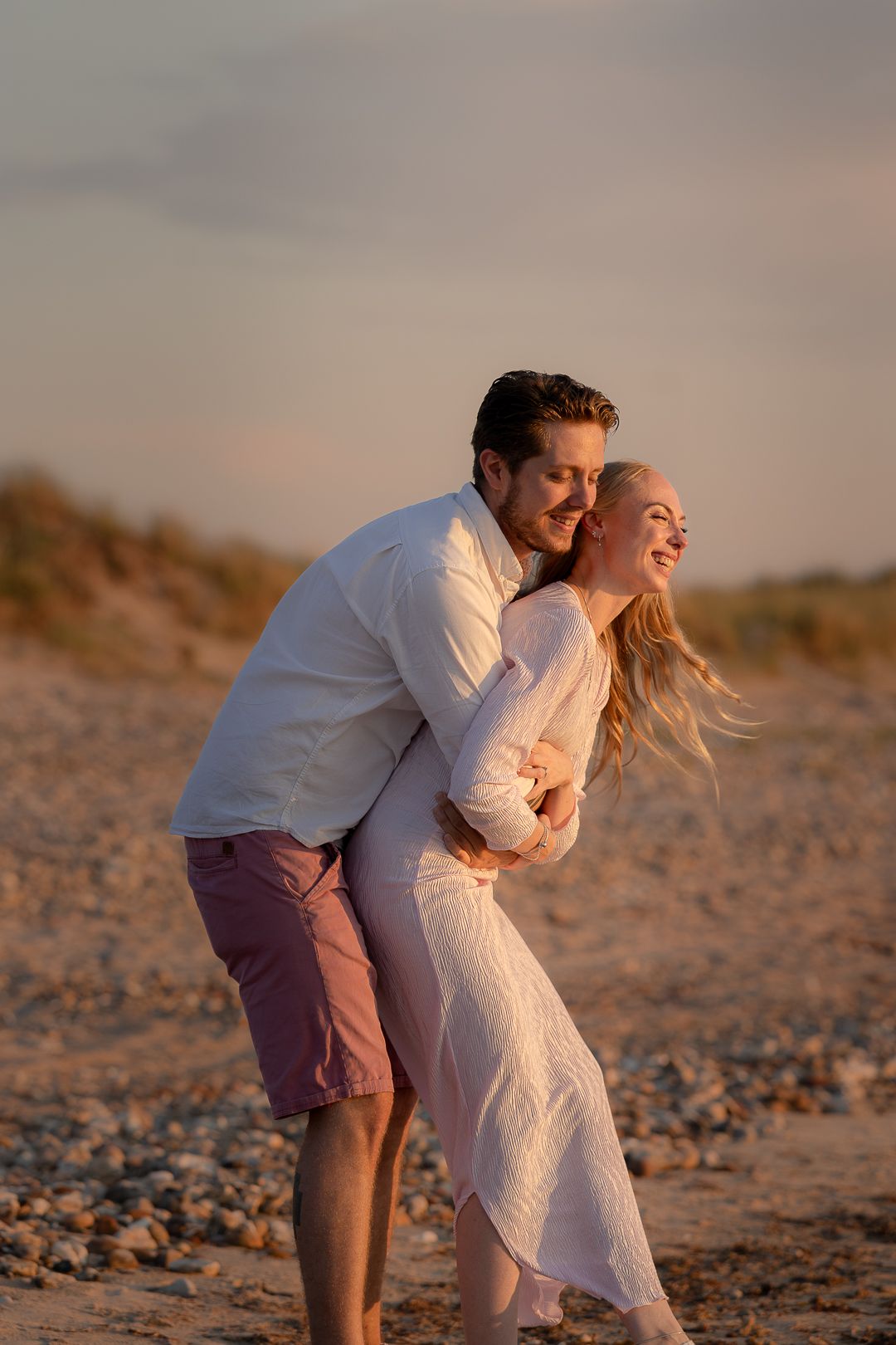 Playful couple laughing and embracing on pebble beach at golden hour sunset.
