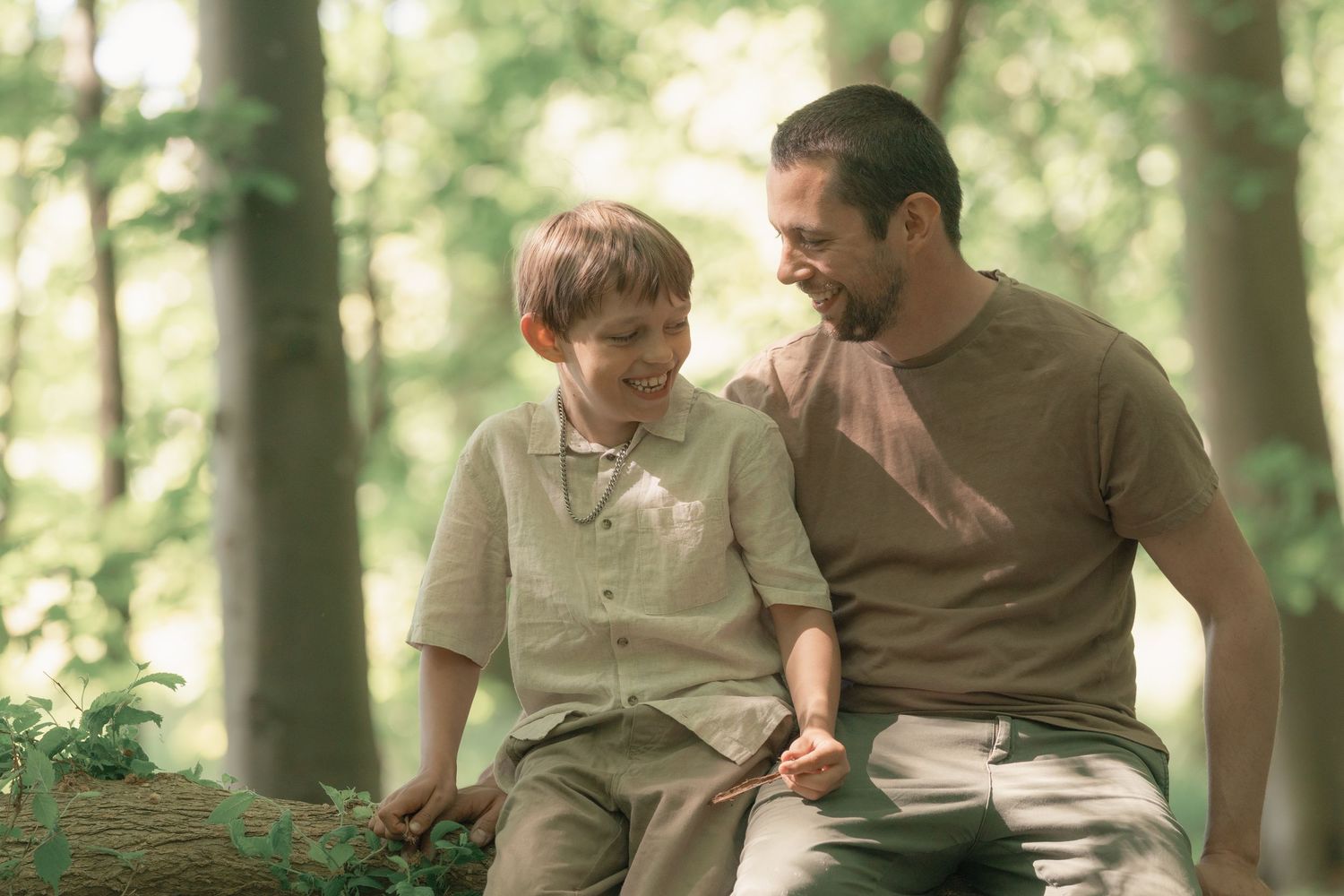 Father and son sitting on log smiling at each other in green forest.