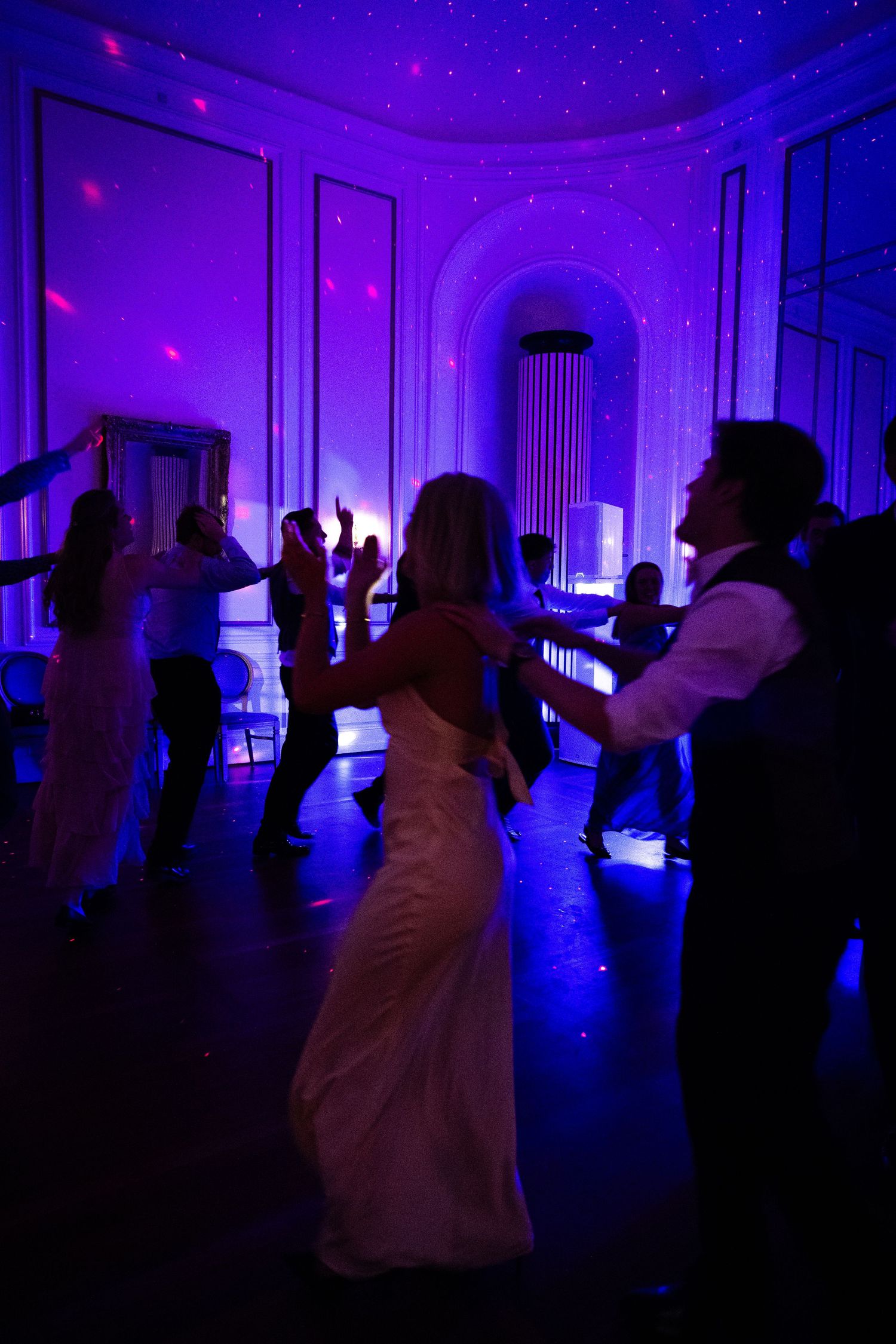 Bride and groom dancing with guests under purple lights at elegant wedding reception.
