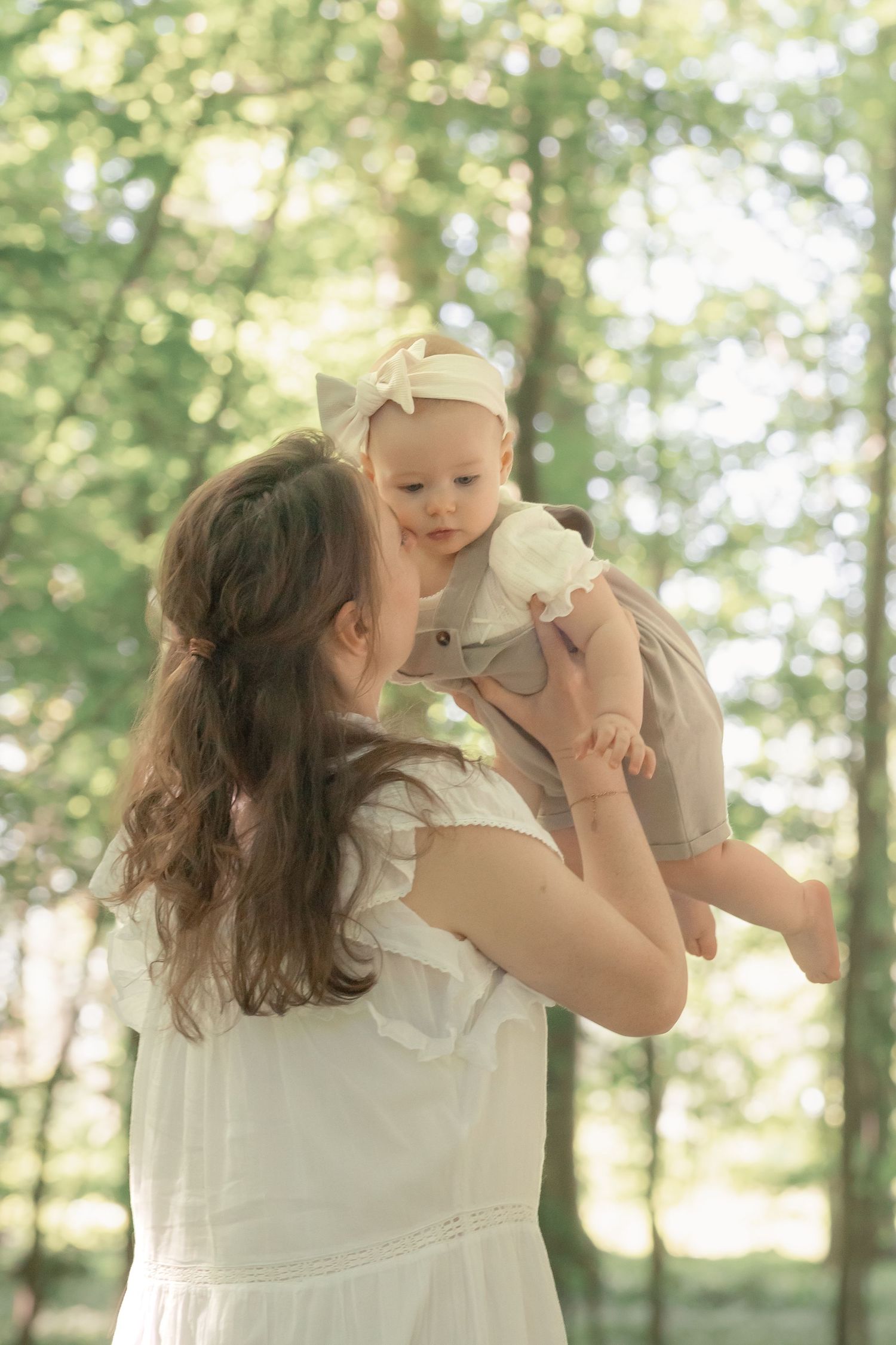 Mother in white dress lifting smiling baby girl with bow headband in sunny forest.