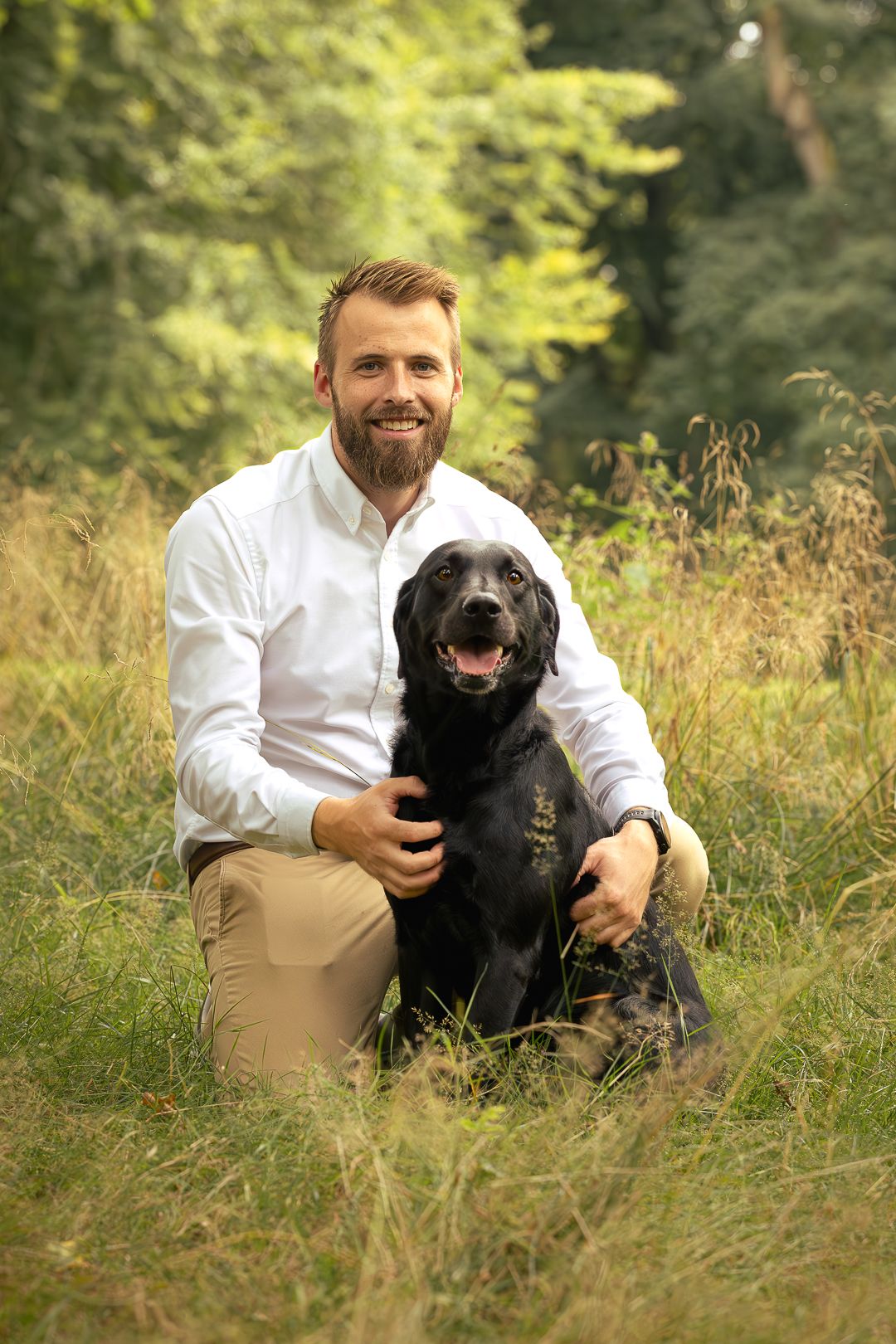 Smiling bearded man sitting with black Labrador dog in green outdoor field.