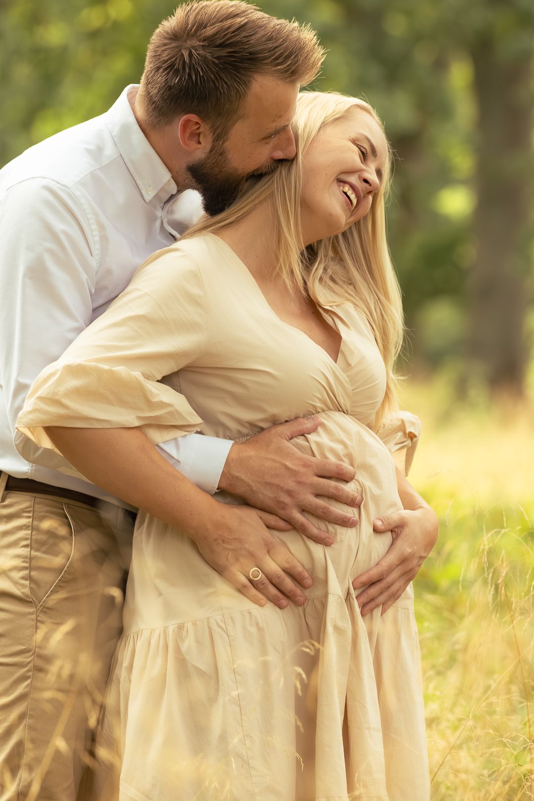 Happy pregnant woman laughing as partner embraces her from behind in sunny golden field.