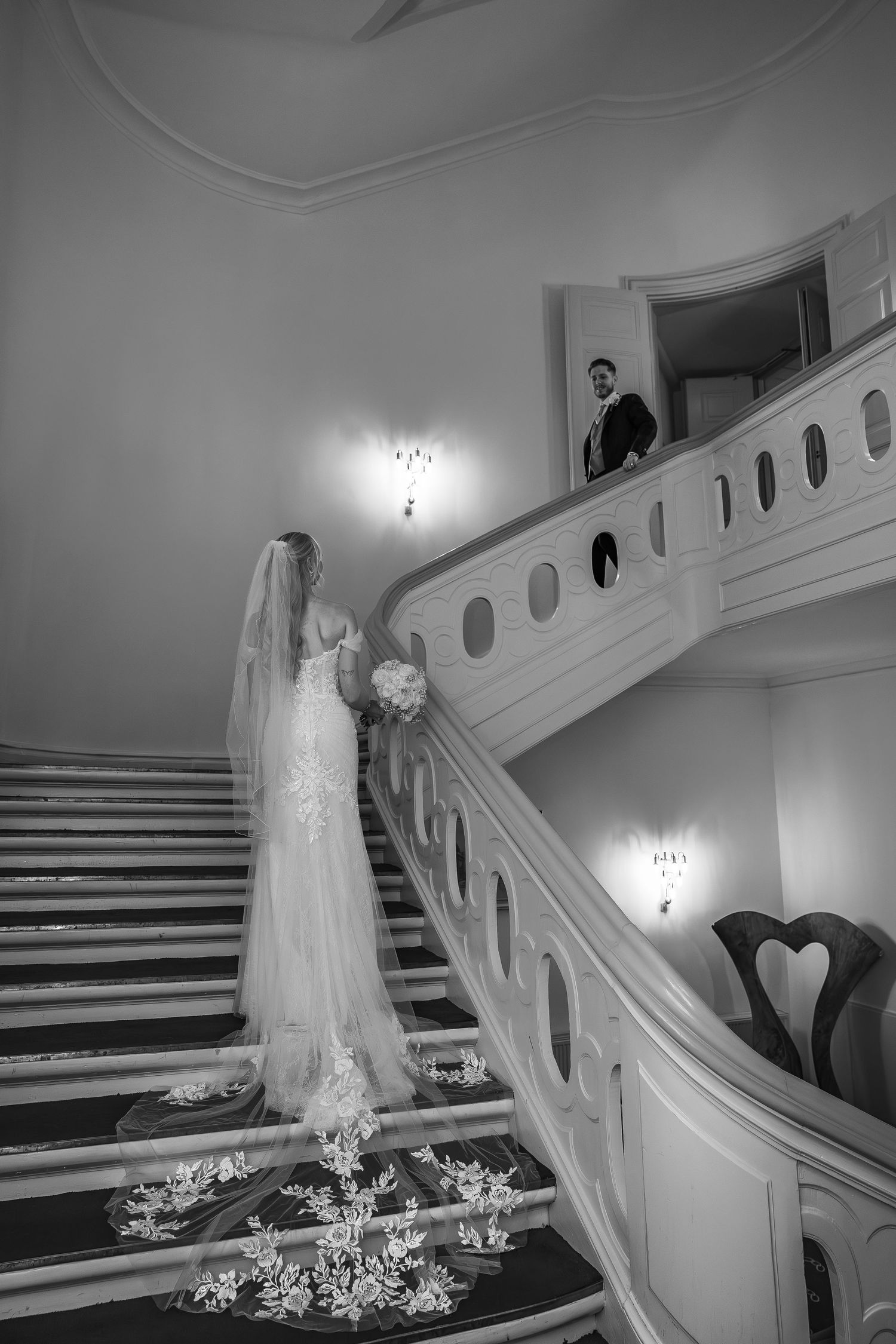 Bride with floral veil on grand staircase as groom watches from above.