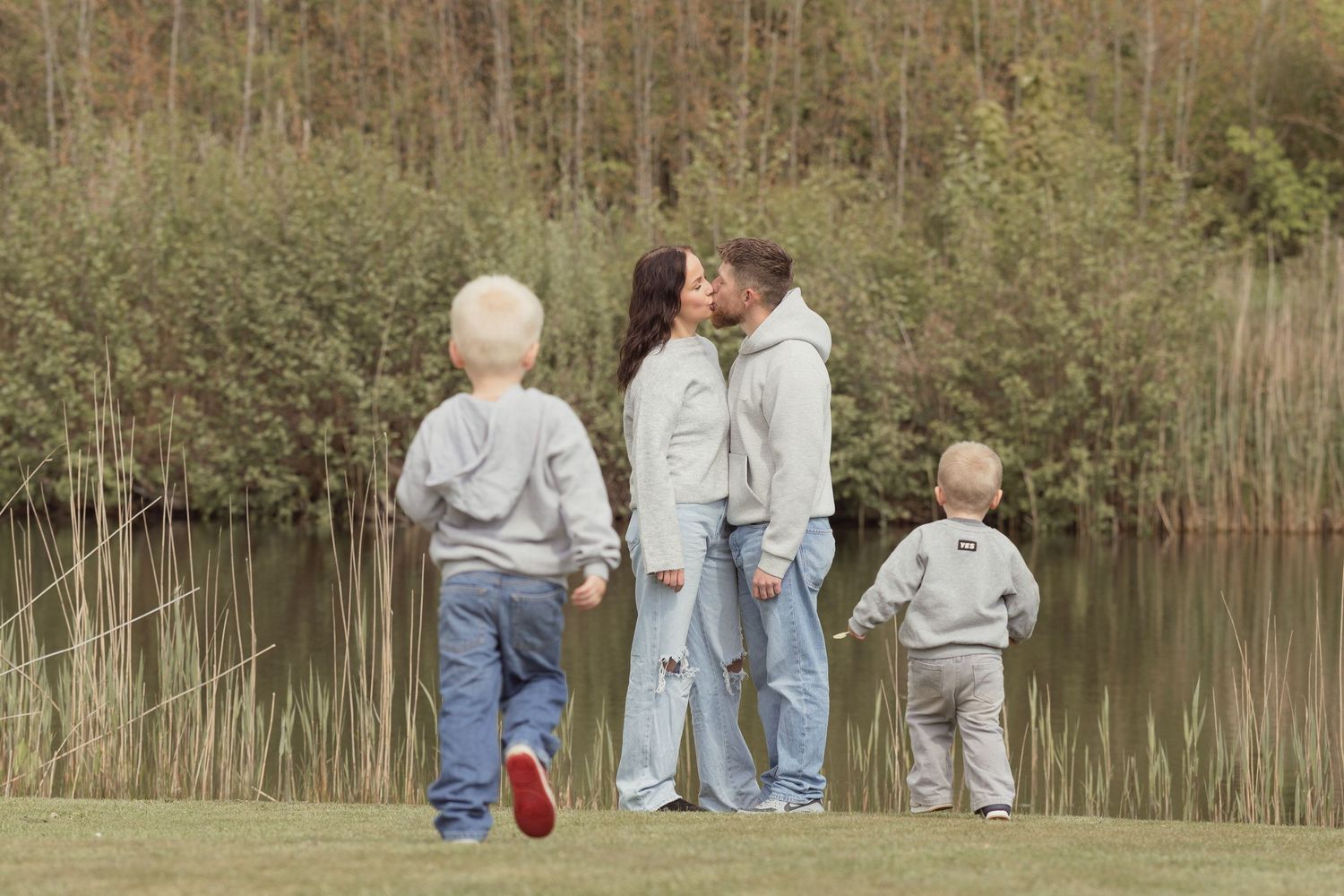 Family of four near a lake, parents kissing while two young boys in matching grey hoodies stand nearby.