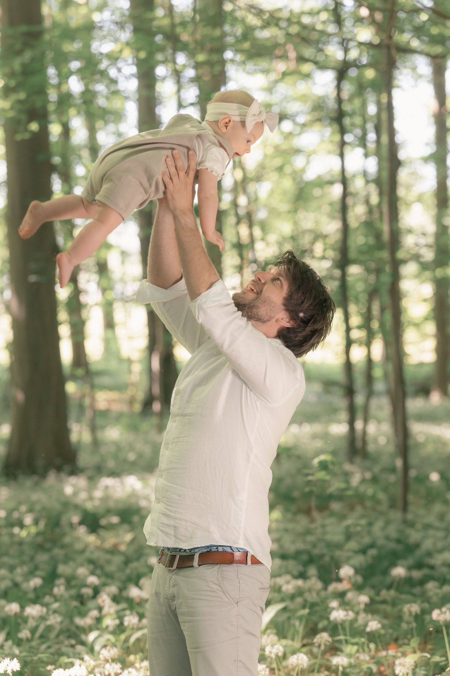 A joyful father raises his baby daughter overhead in a forest filled with wildflowers.