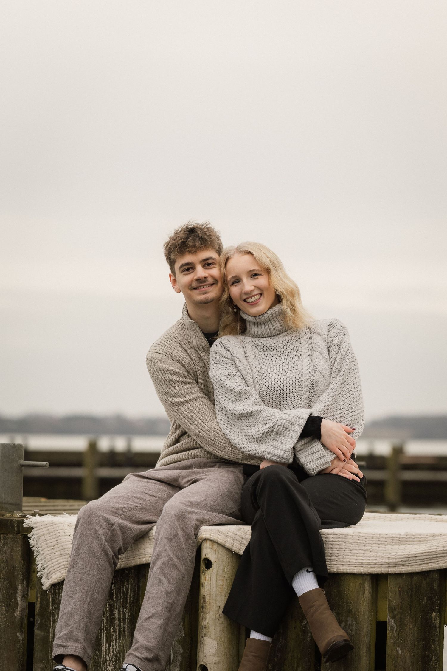 Young couple in matching knit sweaters sitting close together on a dock by the water.