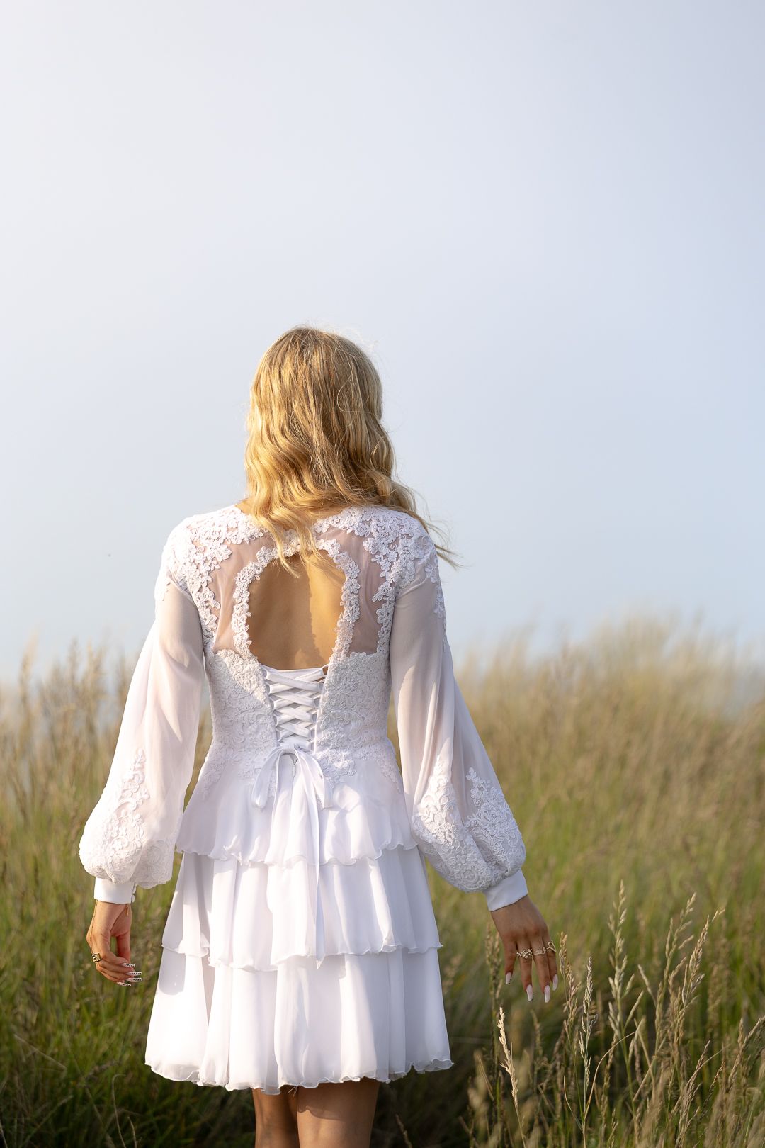 Rear view of blonde girl in white lace confirmation dress walking through tall grass field.