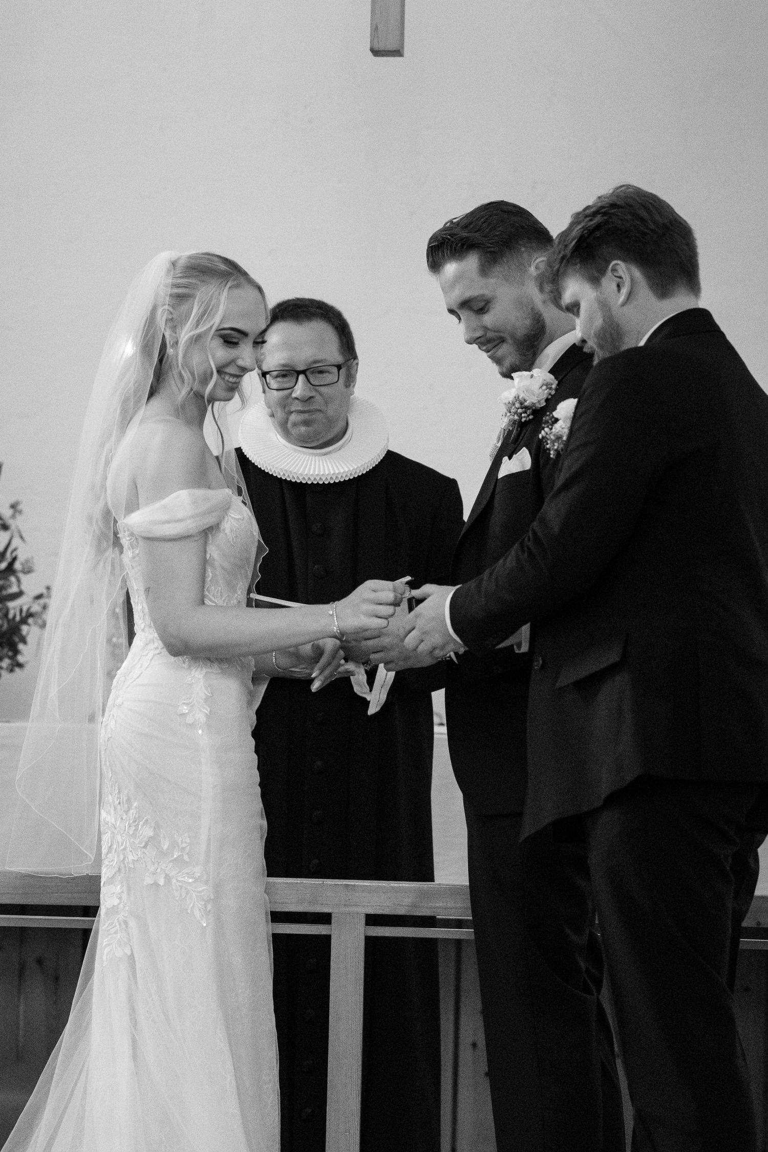 Bride and groom exchanging rings at church altar with officiant in black and white.