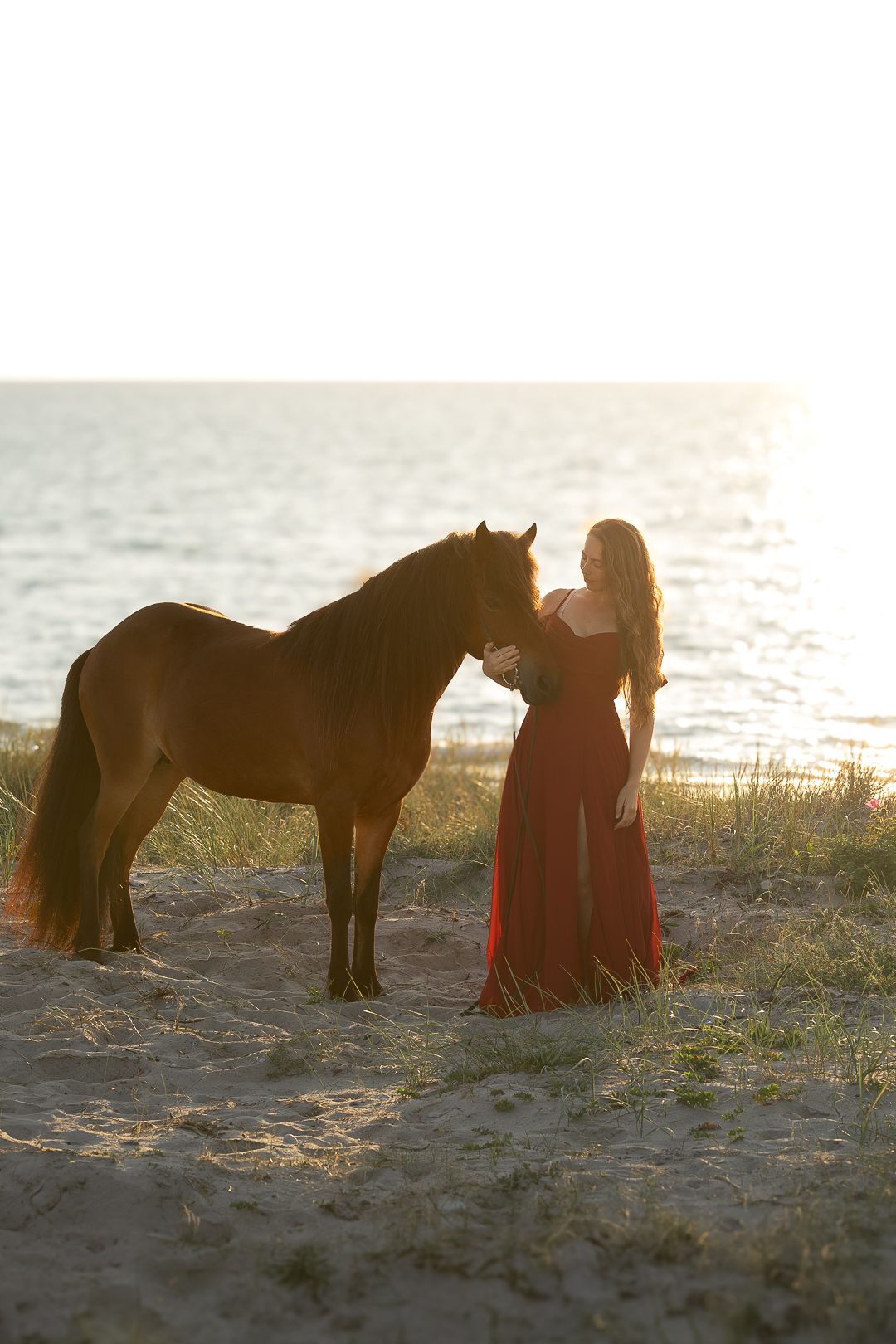 Woman in red dress embracing brown horse on sandy dunes with ocean backdrop at sunset.