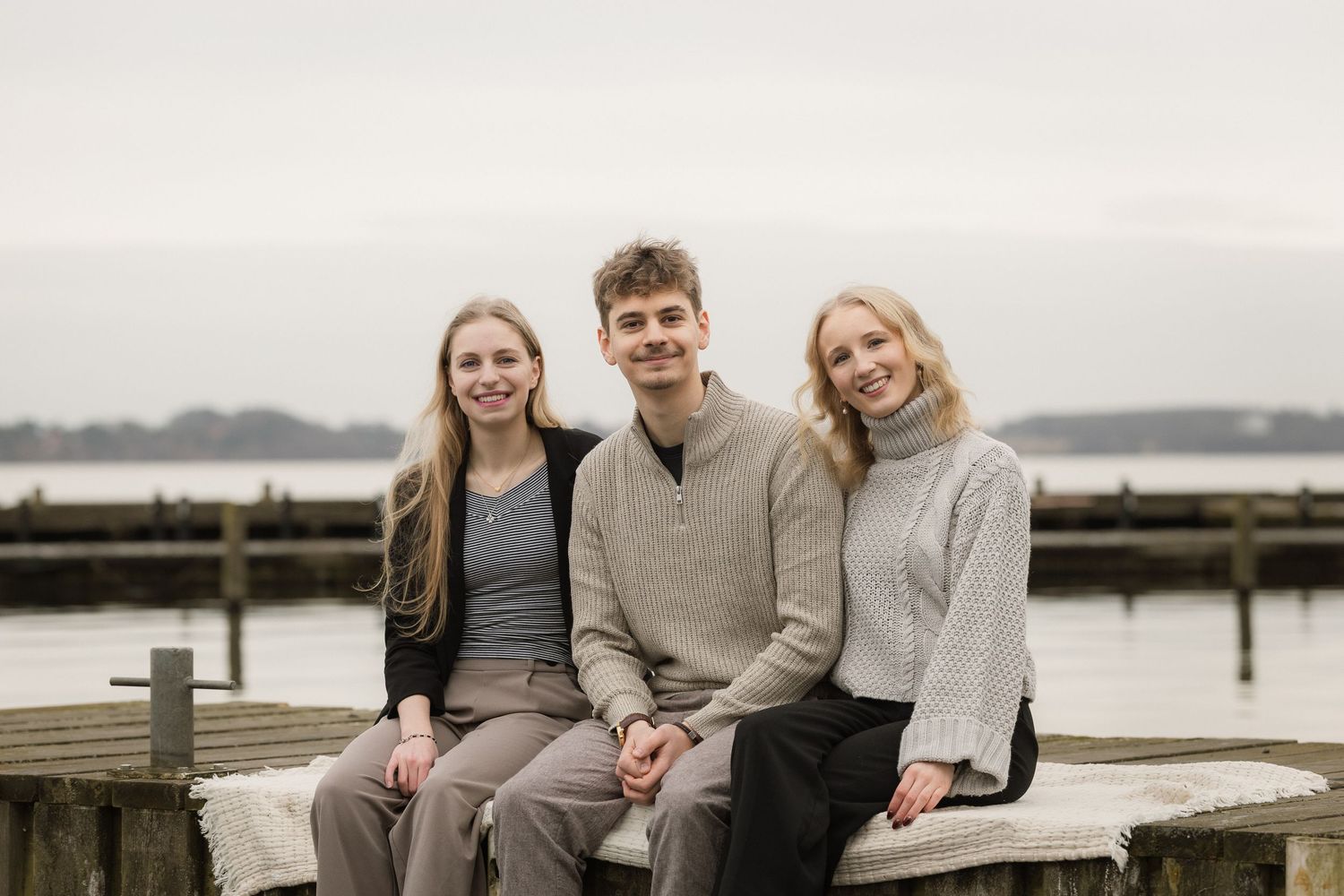 Three teenage siblings sitting together on a dock with calm water behind them.