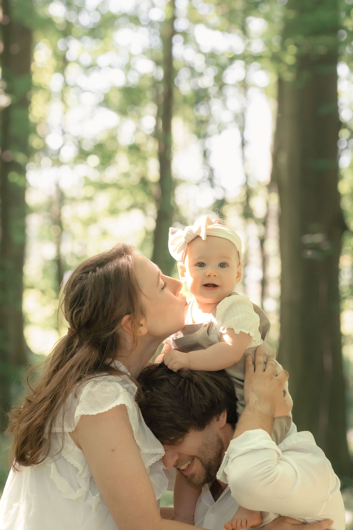 A mother kisses her baby while father bows his head in a sunlit forest family portrait.