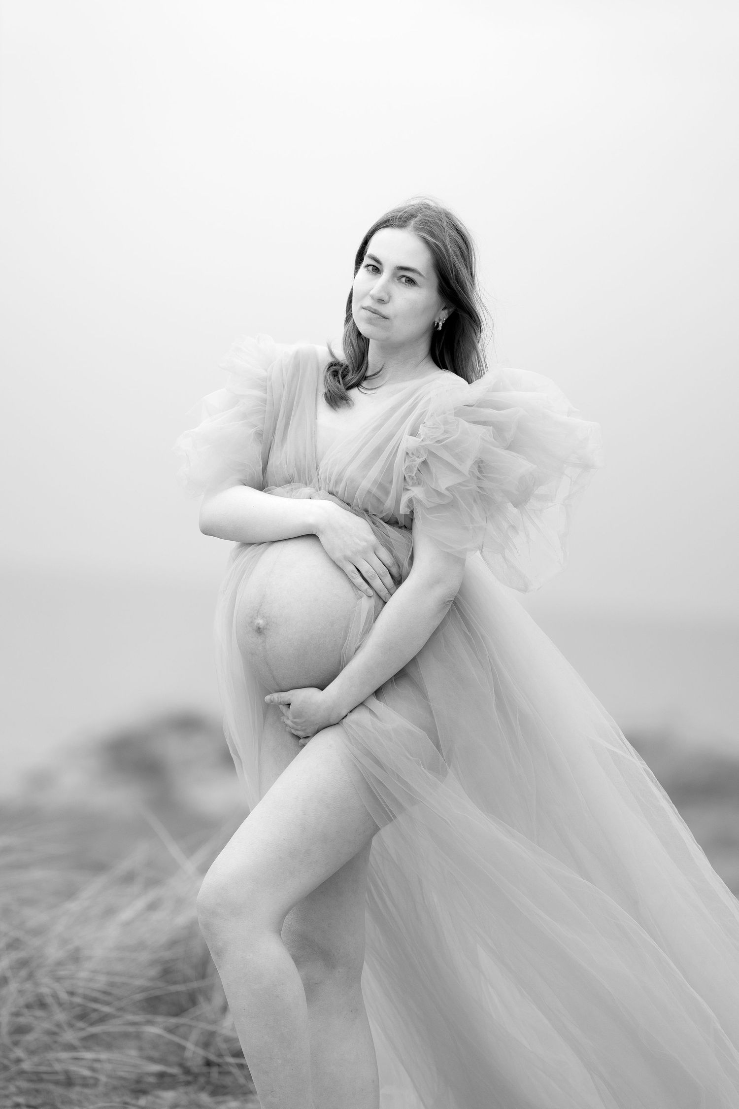 Black and white portrait of pregnant woman in sheer tulle gown embracing her baby bump by the sea.