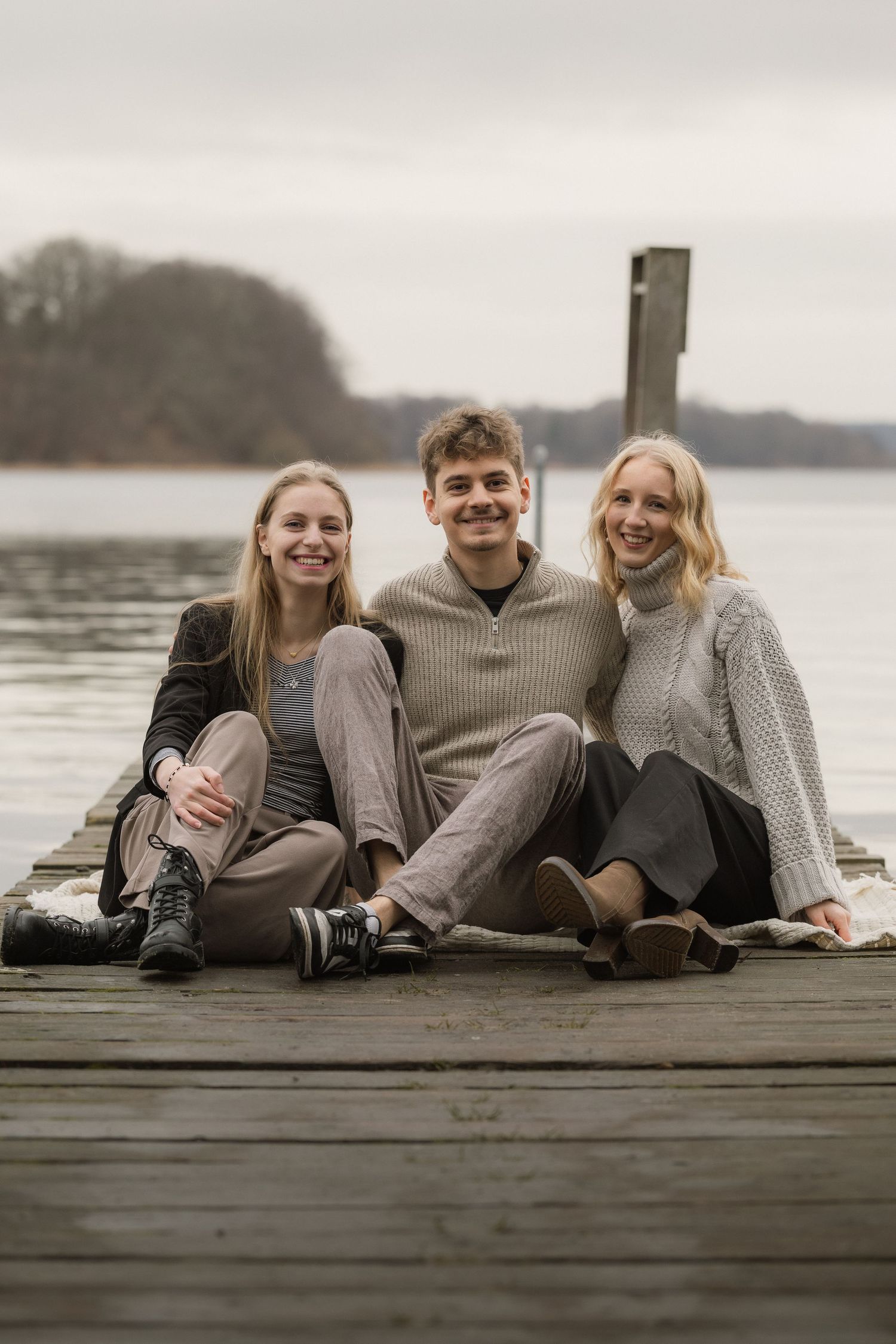 Three smiling young adults sit together on a wooden dock by a calm lake on an overcast day.