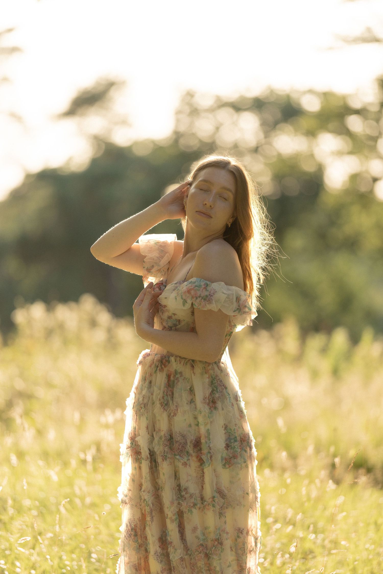 Young woman in floral off-shoulder dress standing in sunlit meadow with golden backlight and trees.