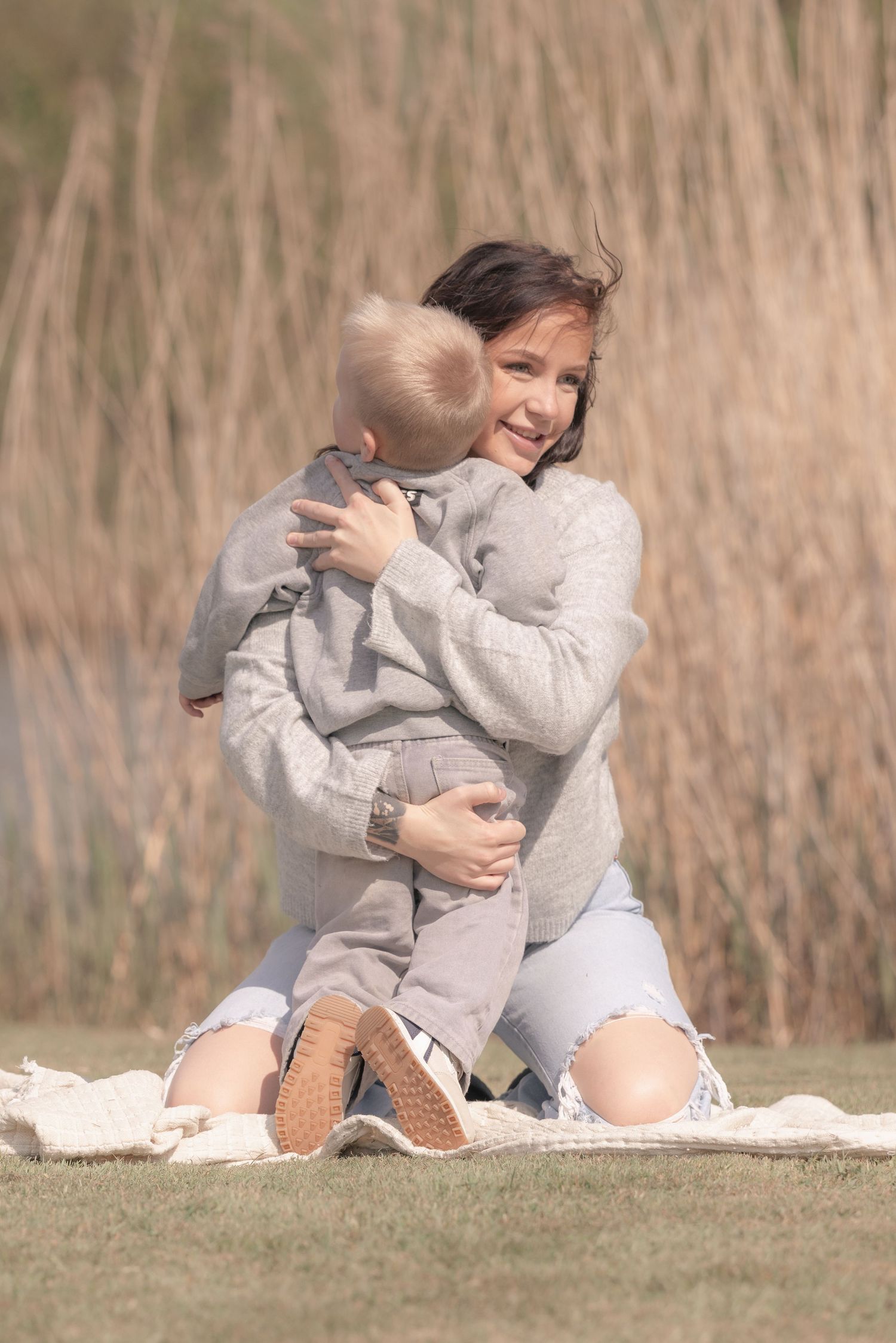 Smiling mother hugging her young blonde son while sitting on a blanket outdoors in nature.