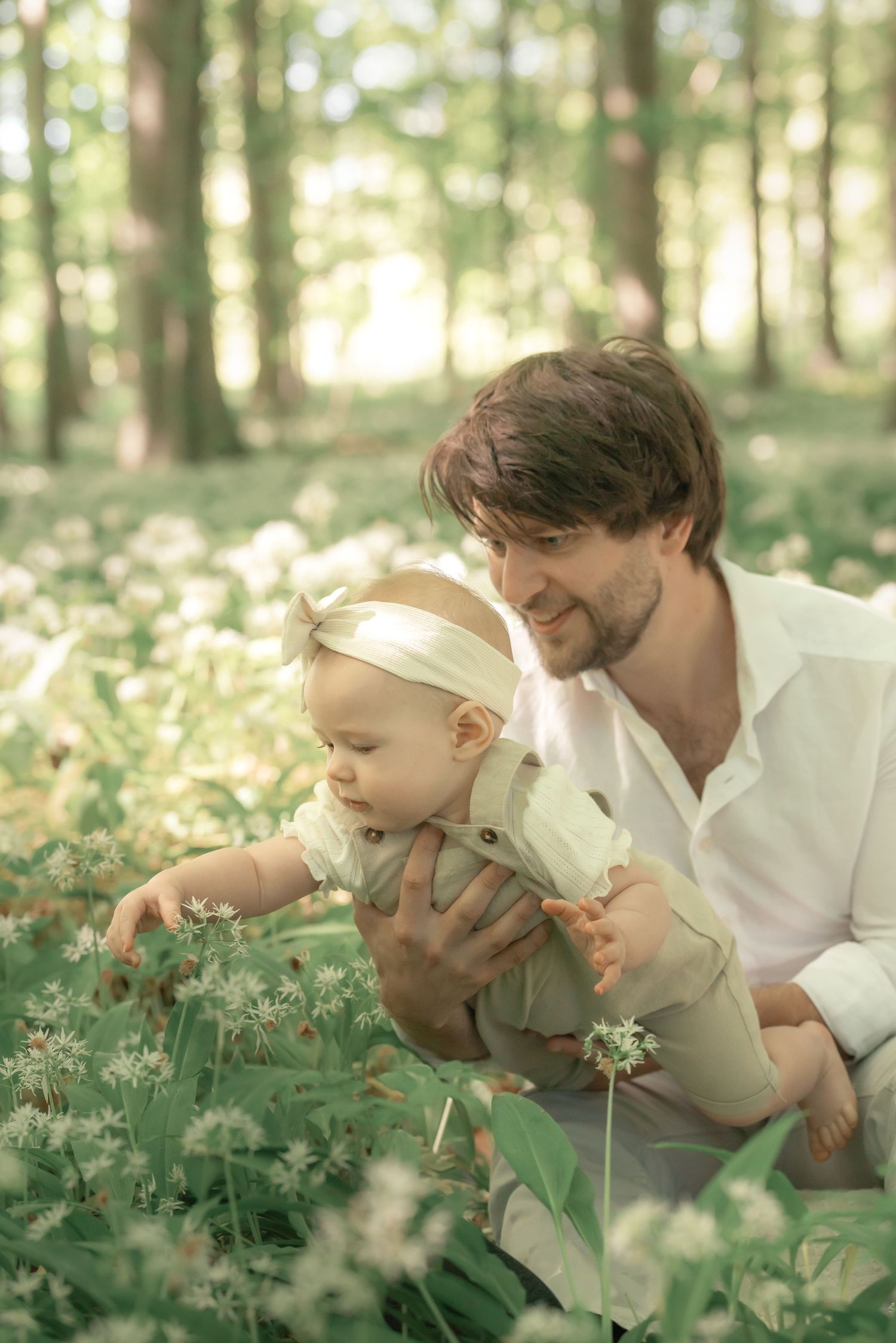 A smiling father helps his curious baby explore white wildflowers in a green forest.
