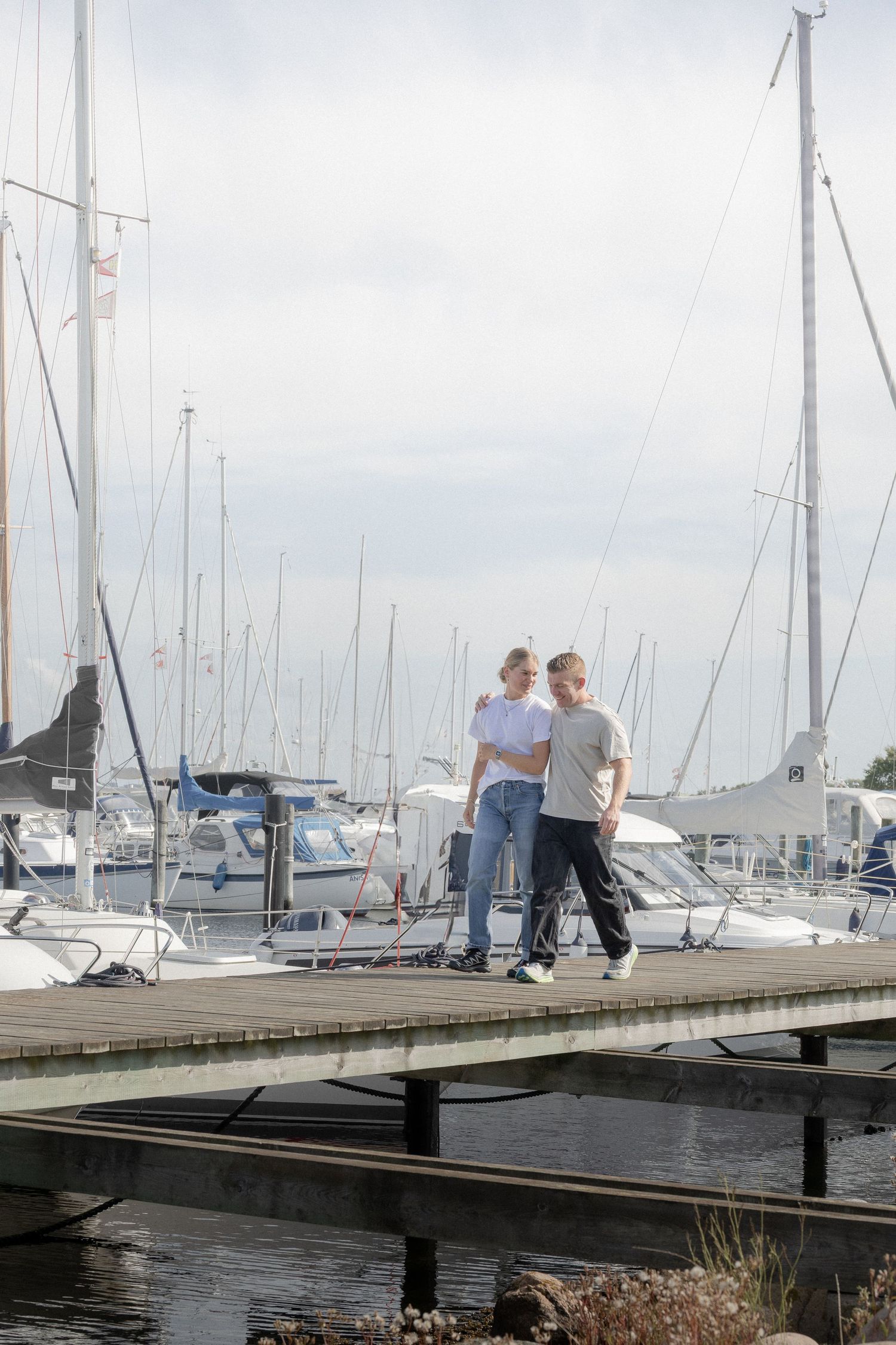 A young couple walks hand in hand on a wooden marina dock surrounded by sailboats on a sunny day.