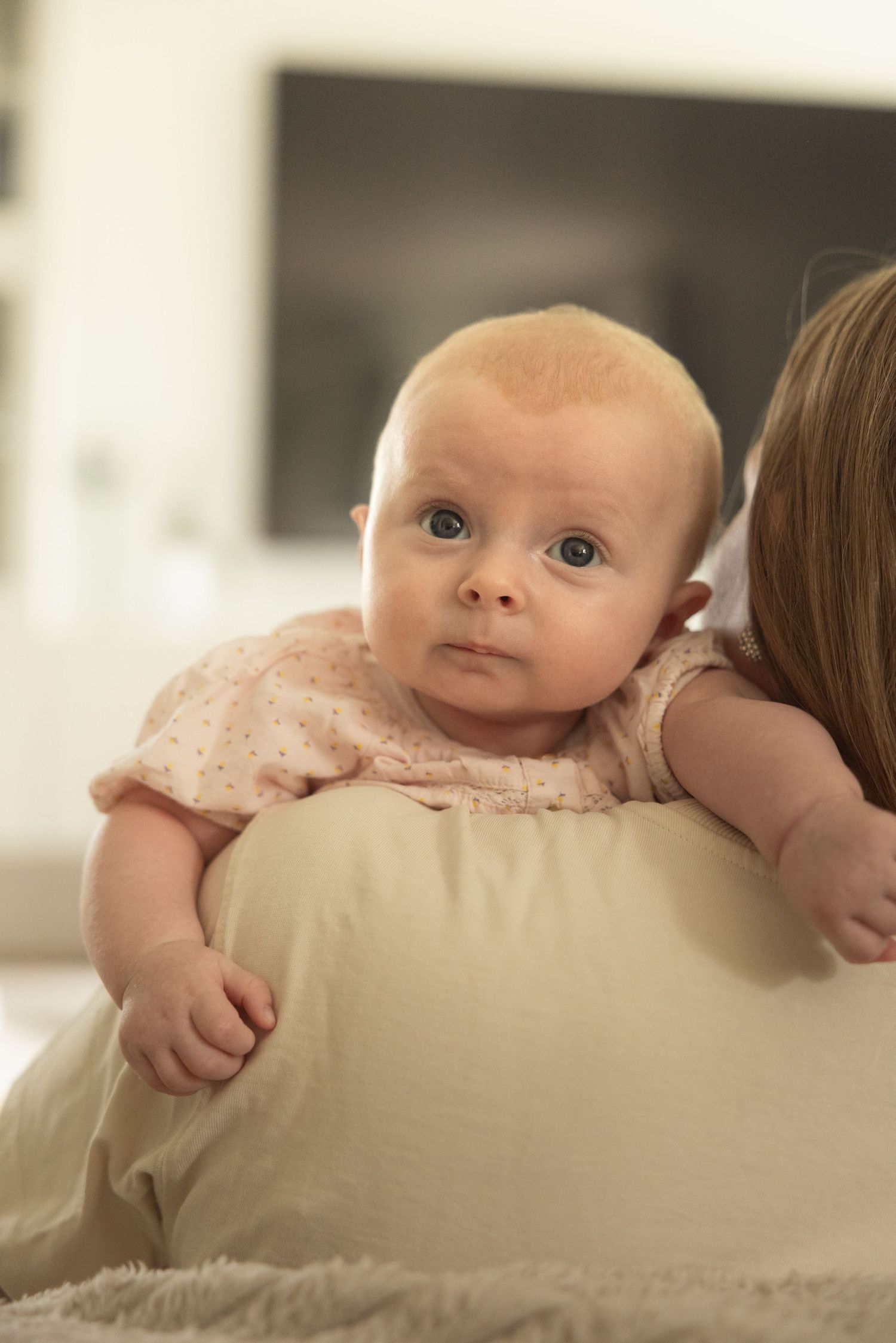 A curious blue-eyed baby in a pink outfit rests on a mother's shoulder, looking directly at the camera.
