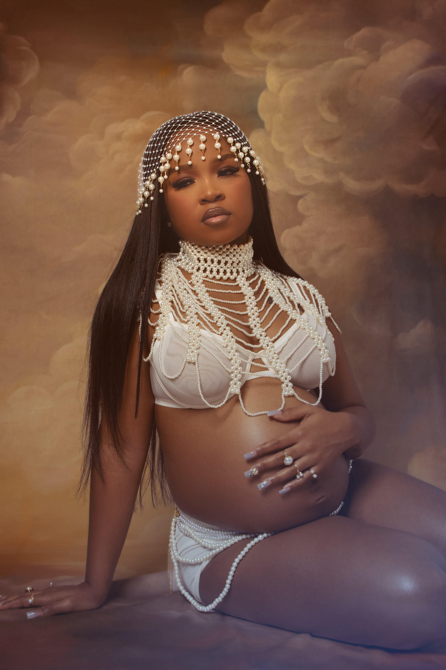 Pregnant woman in white pearl macrame outfit poses seated against dramatic cloudy background.