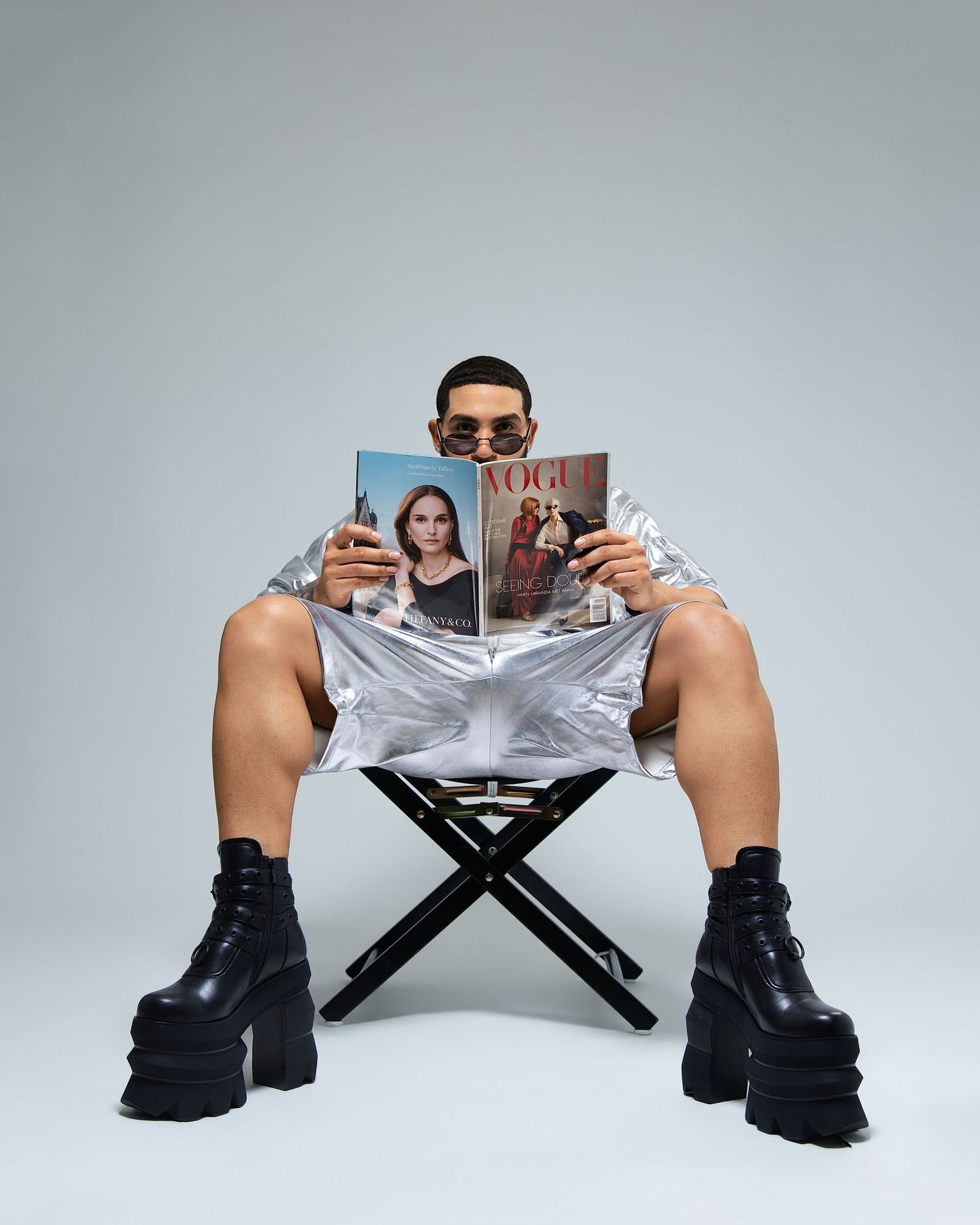 Man in silver metallic outfit reading Vogue magazine while seated on folding chair with platform boots.