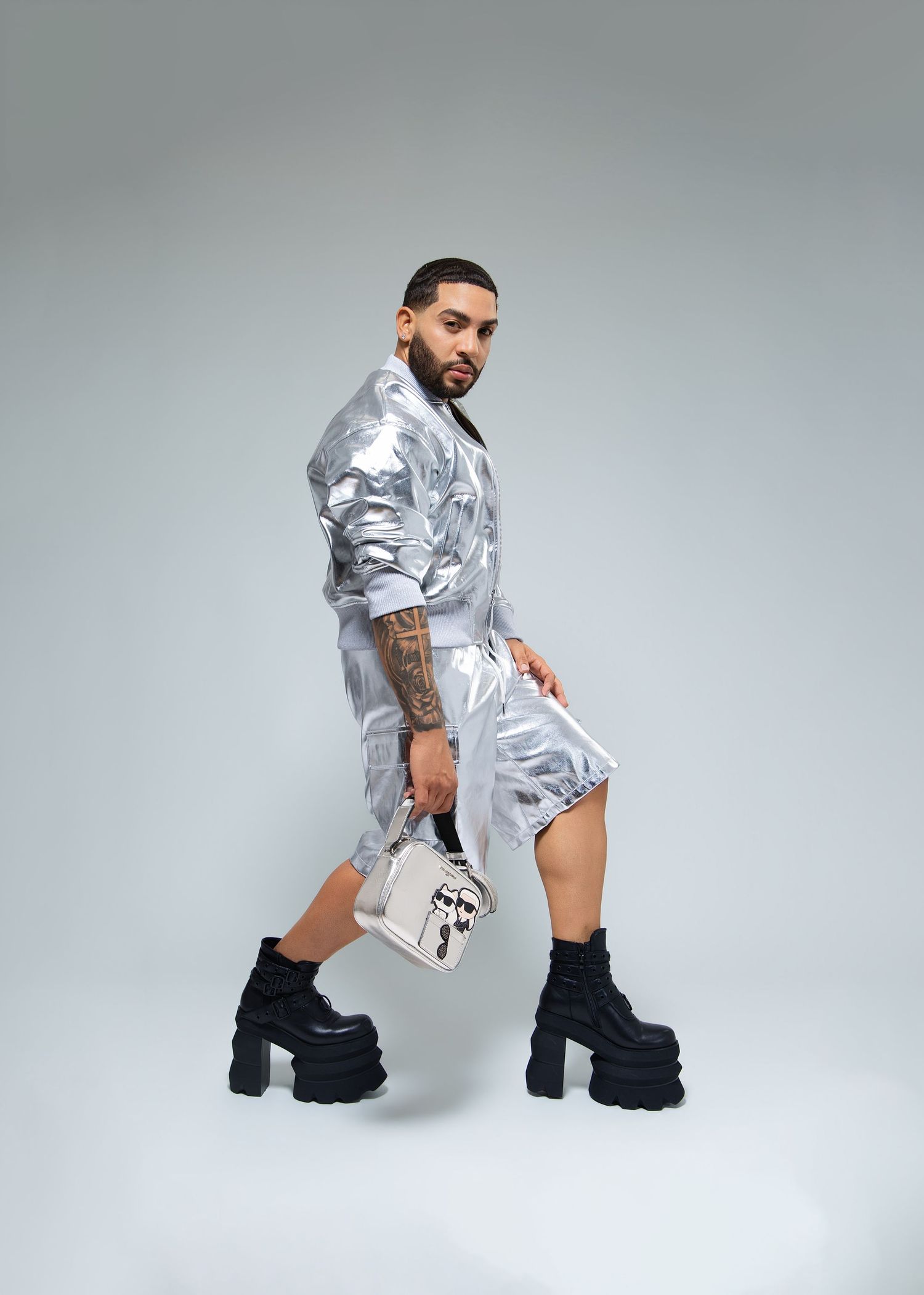 Man in silver metallic jacket and shorts holding white handbag wearing black platform boots, side profile.