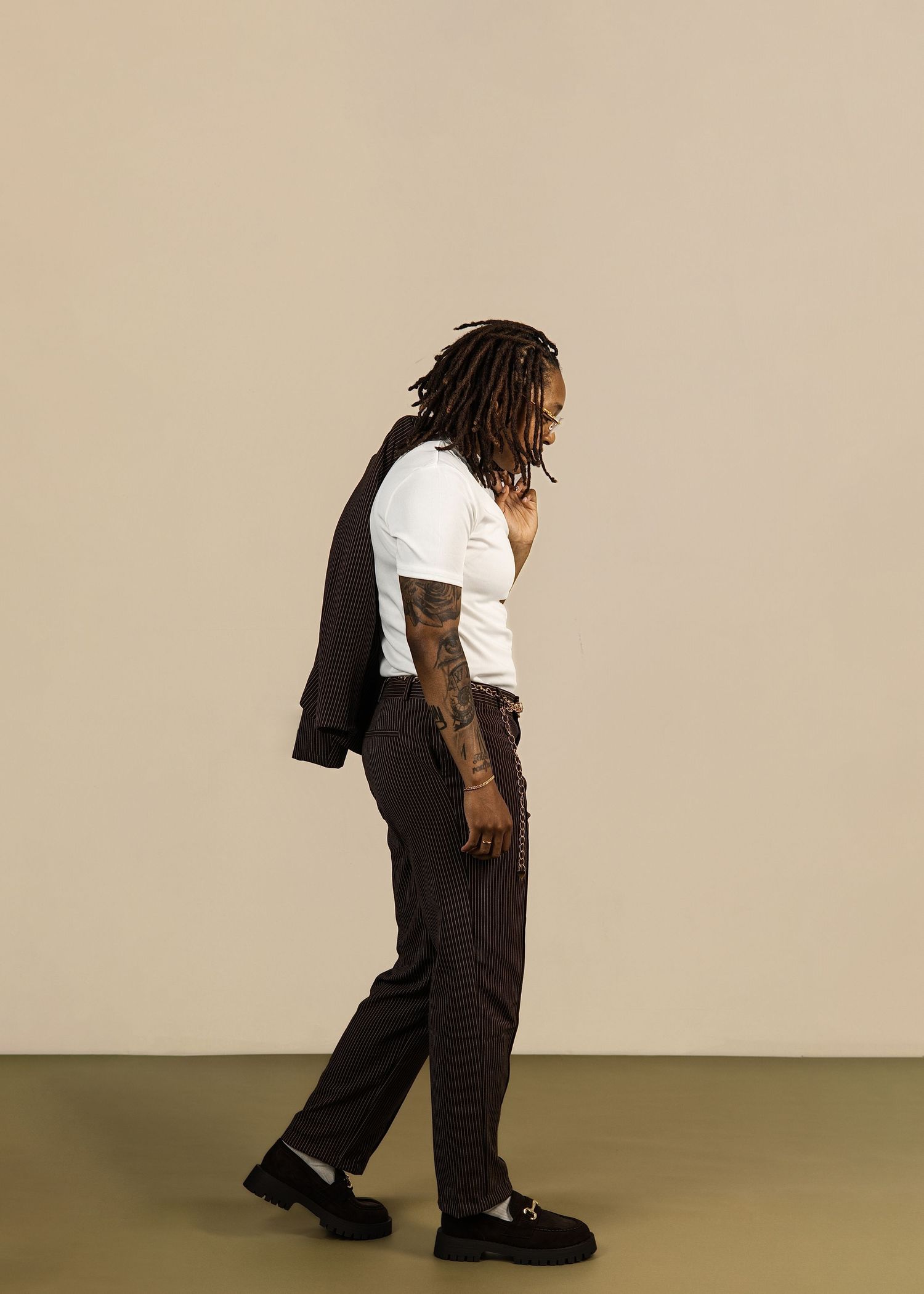 Person with locs and tattoos walks in profile view against neutral studio background.