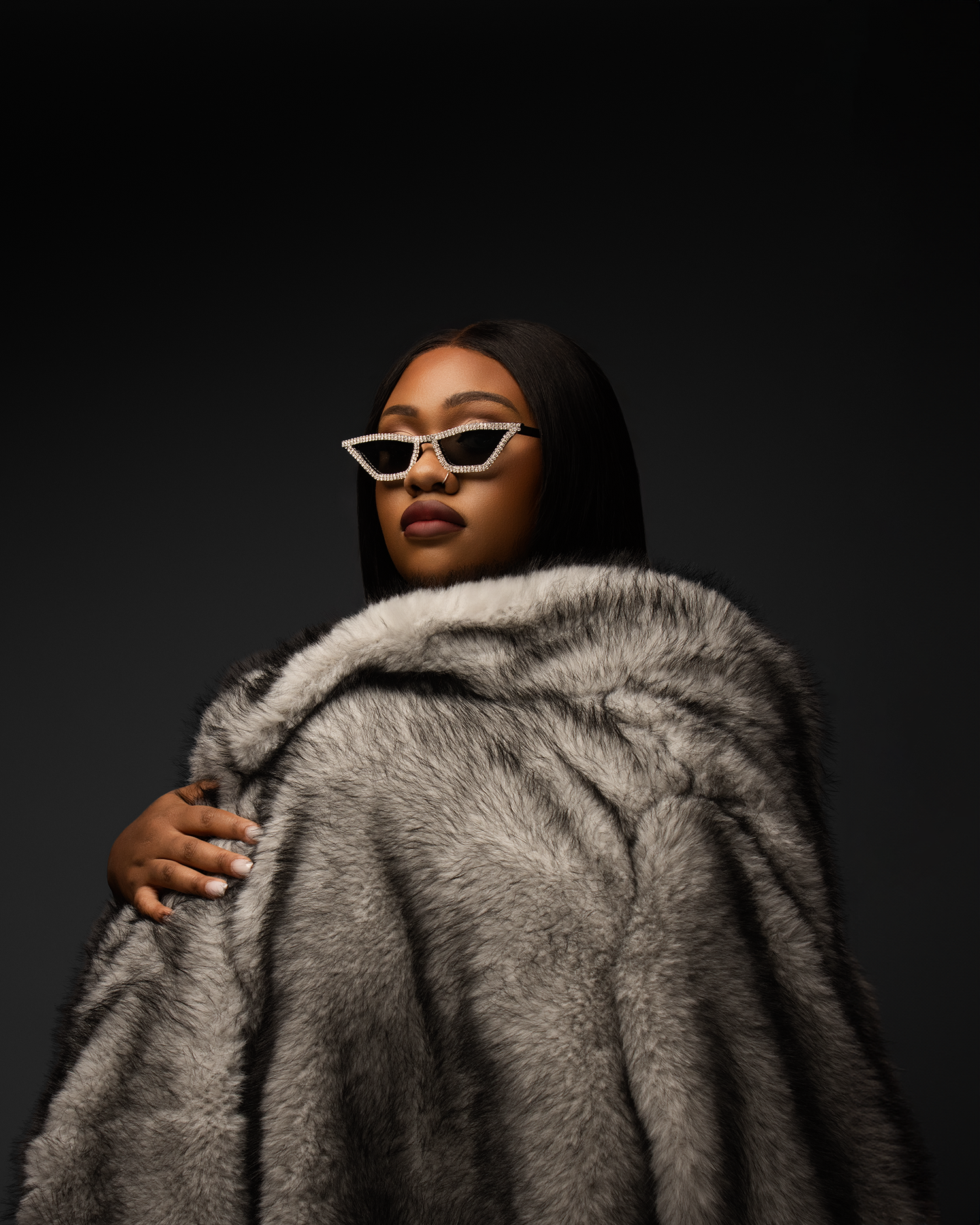 Woman in gray faux fur coat and cat-eye sunglasses poses in dark studio setting.