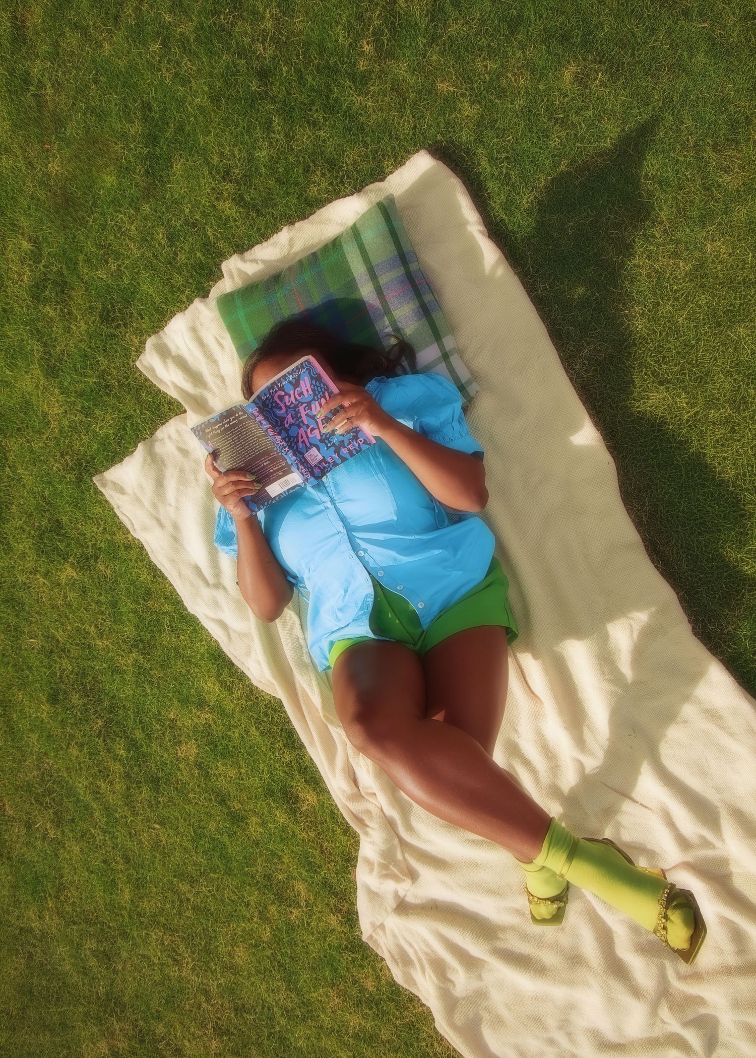 Woman in blue top and green shorts lying on a blanket in grass reading a colorful book outdoors.