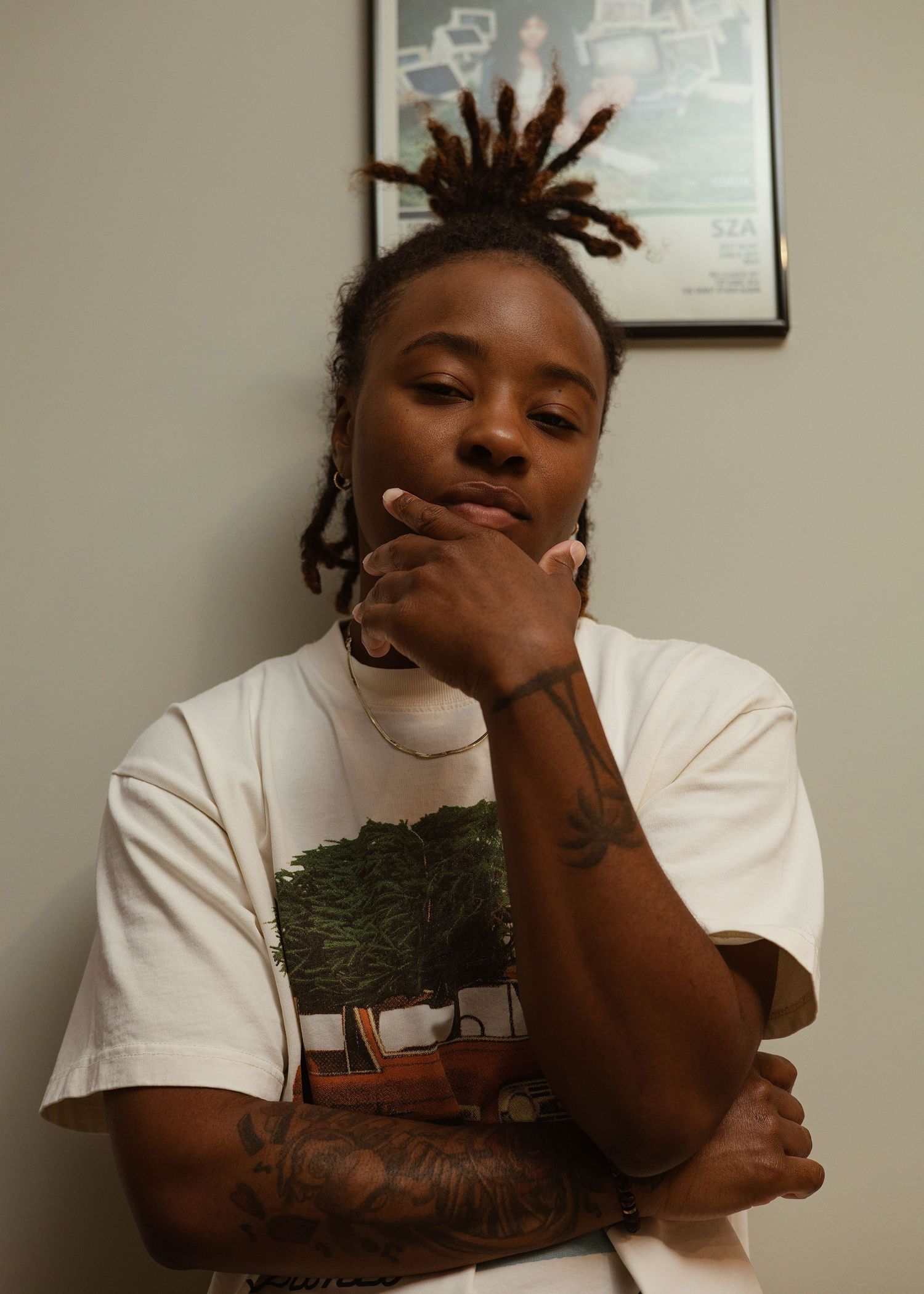 Young Black person with locs and tattoos posing thoughtfully in white graphic tee.