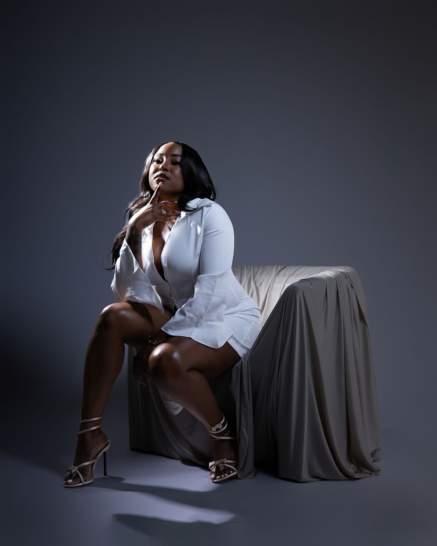 Confident woman in white shirt dress sits on draped chair in dramatic studio lighting.