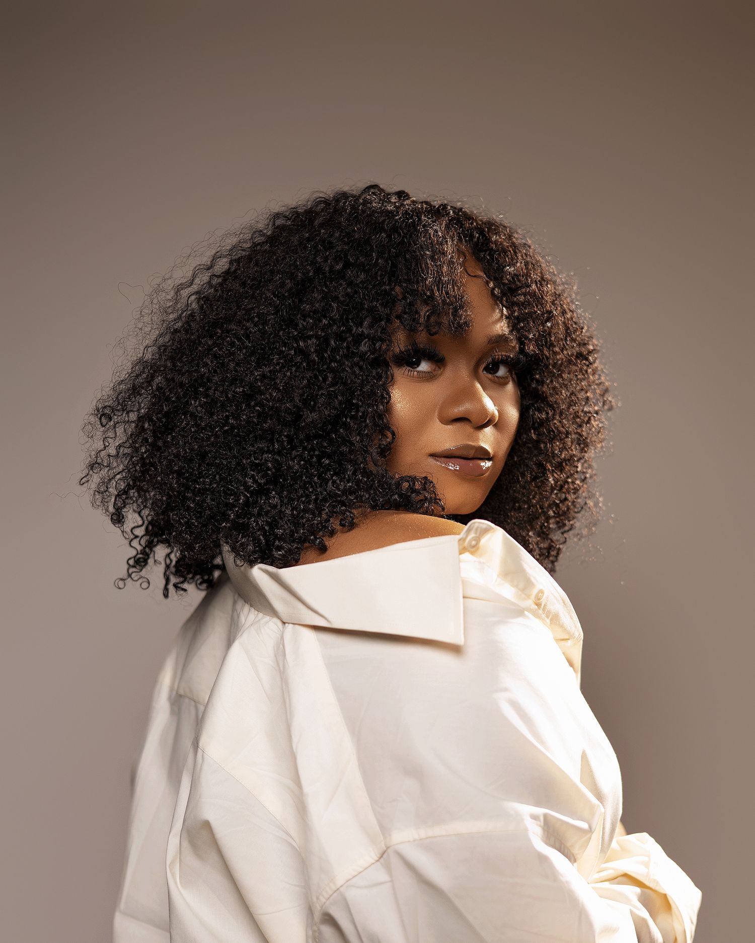 Woman with voluminous curly hair in white off-shoulder shirt posing against a warm gray background.