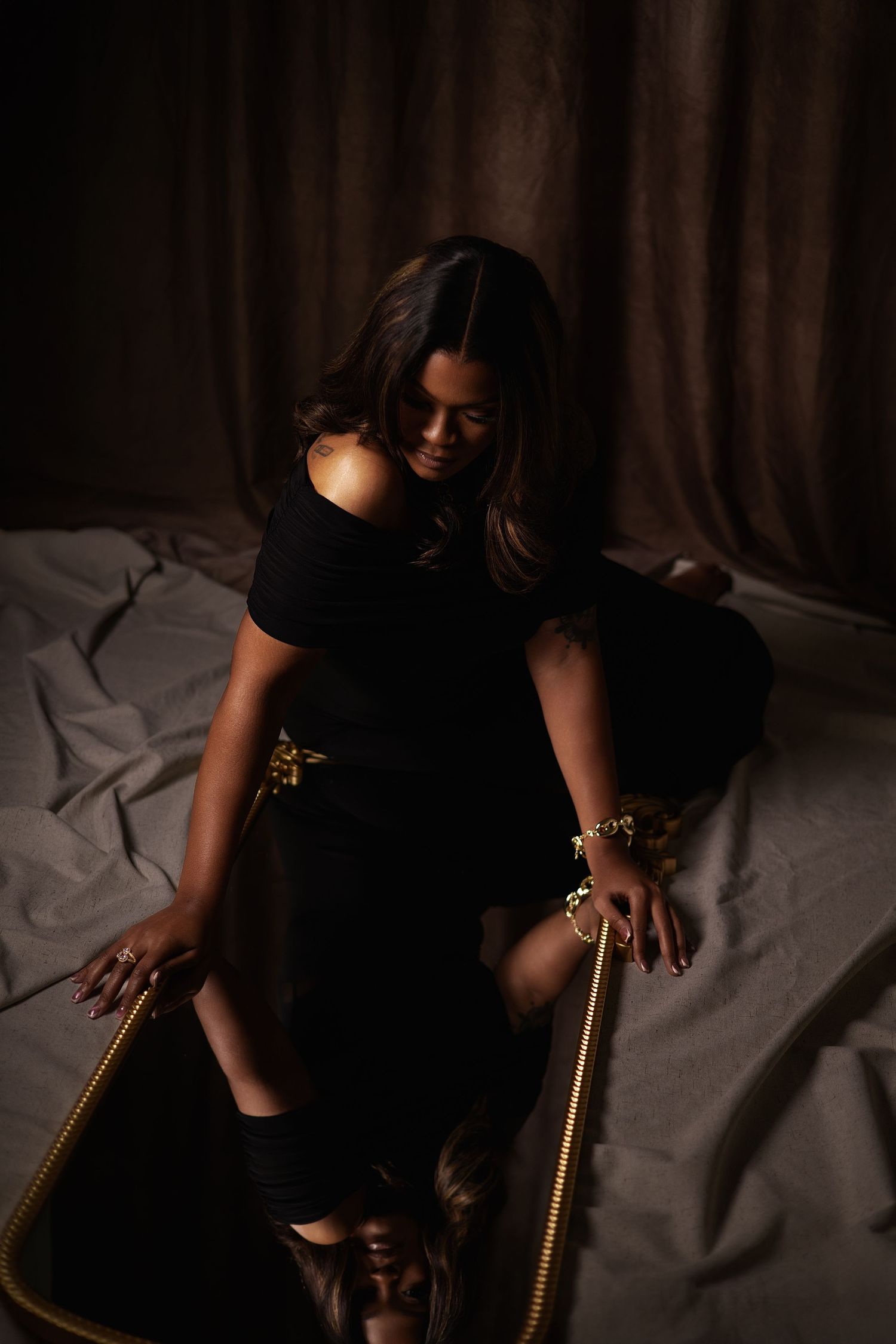 Woman in black off-shoulder dress seated on gold-framed mirror in dramatic moody studio setting.