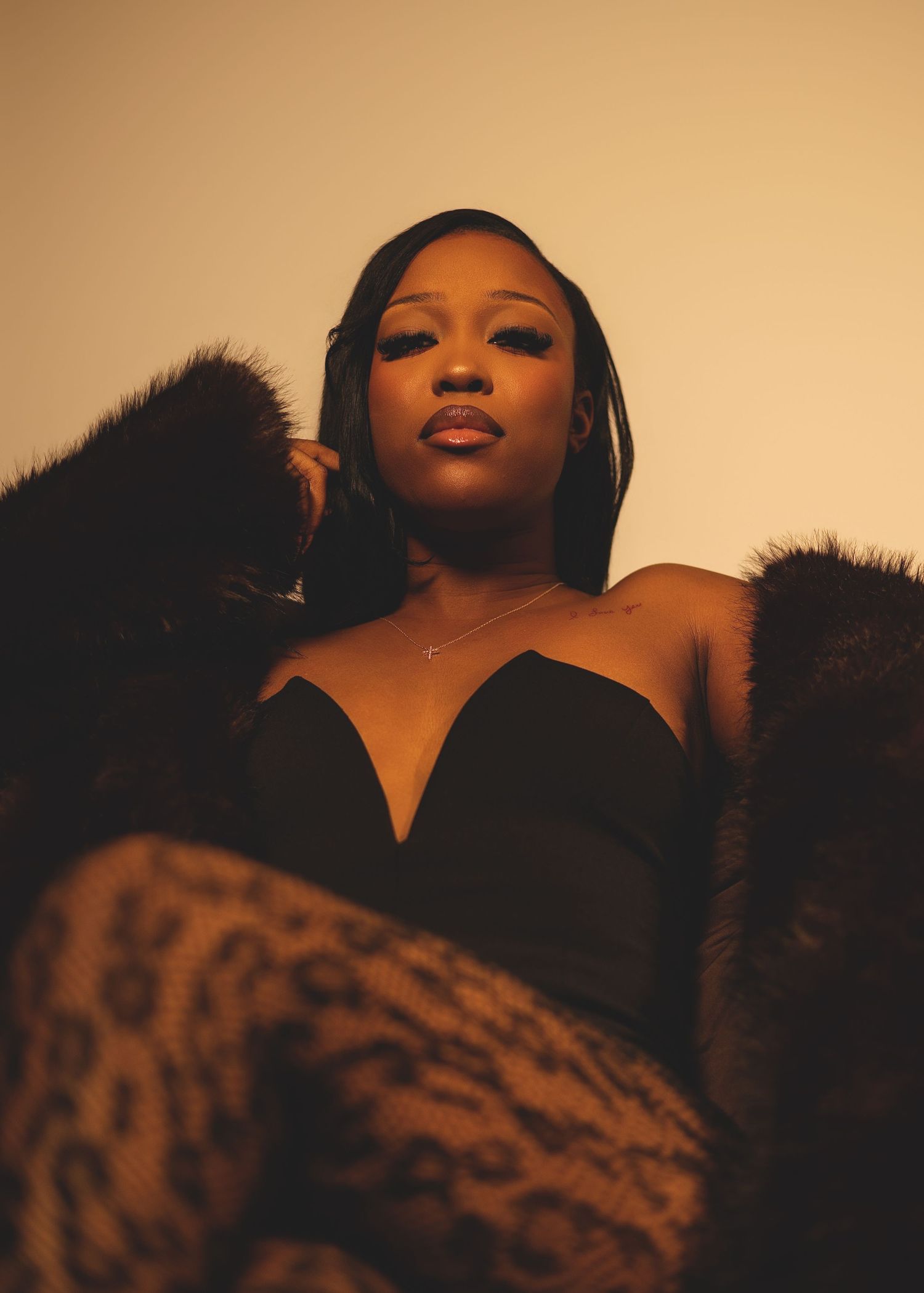 A woman in a black strapless top with a fur wrap gazes upward in warm golden studio lighting.