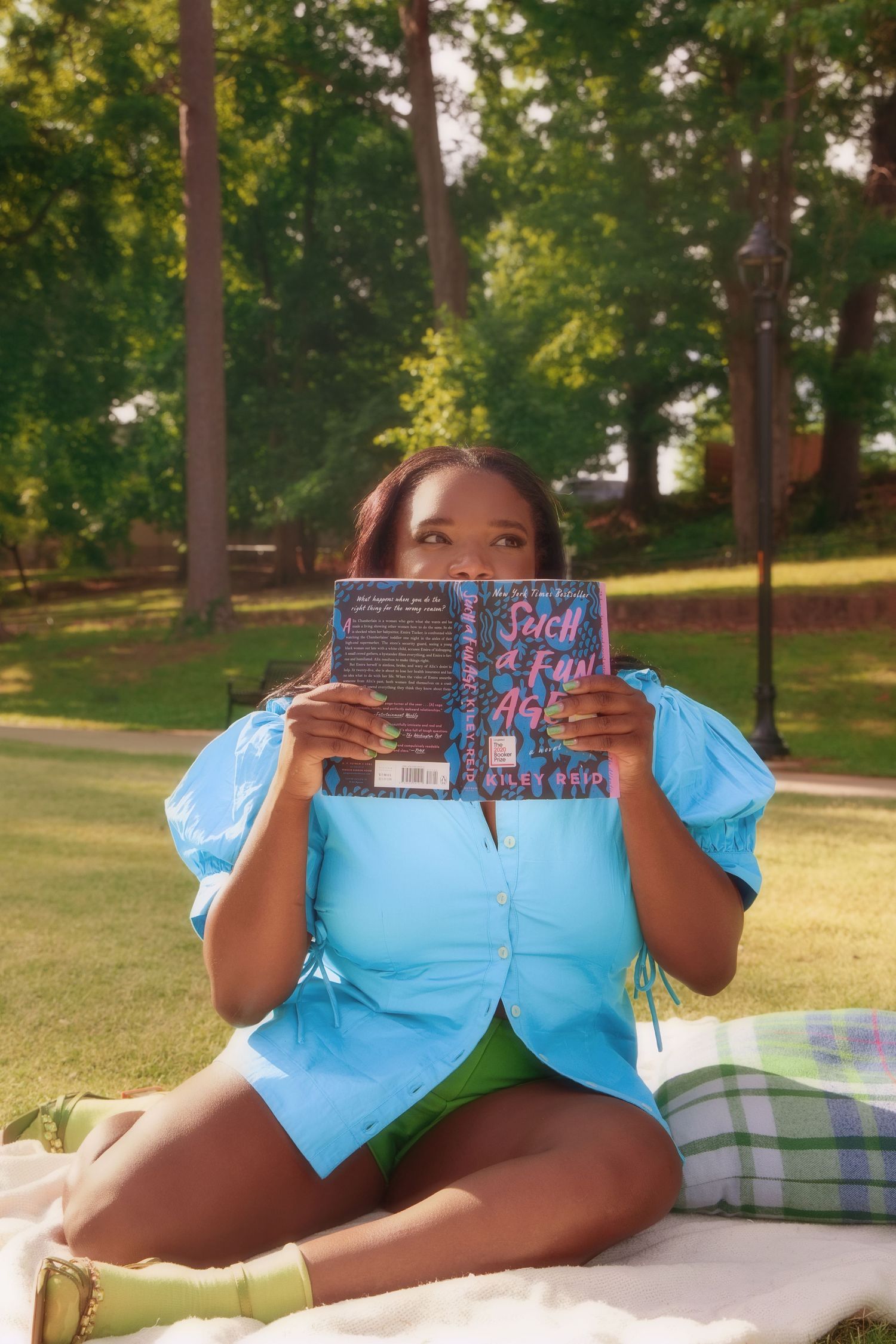 Woman in blue puff-sleeve top and green shorts sitting on blanket in park holding up a colorful book.