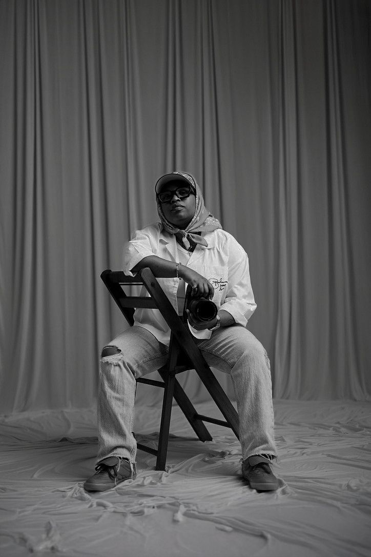 Black and white portrait of a person sitting on a folding chair, wearing sunglasses and a headscarf, holding a camera.
