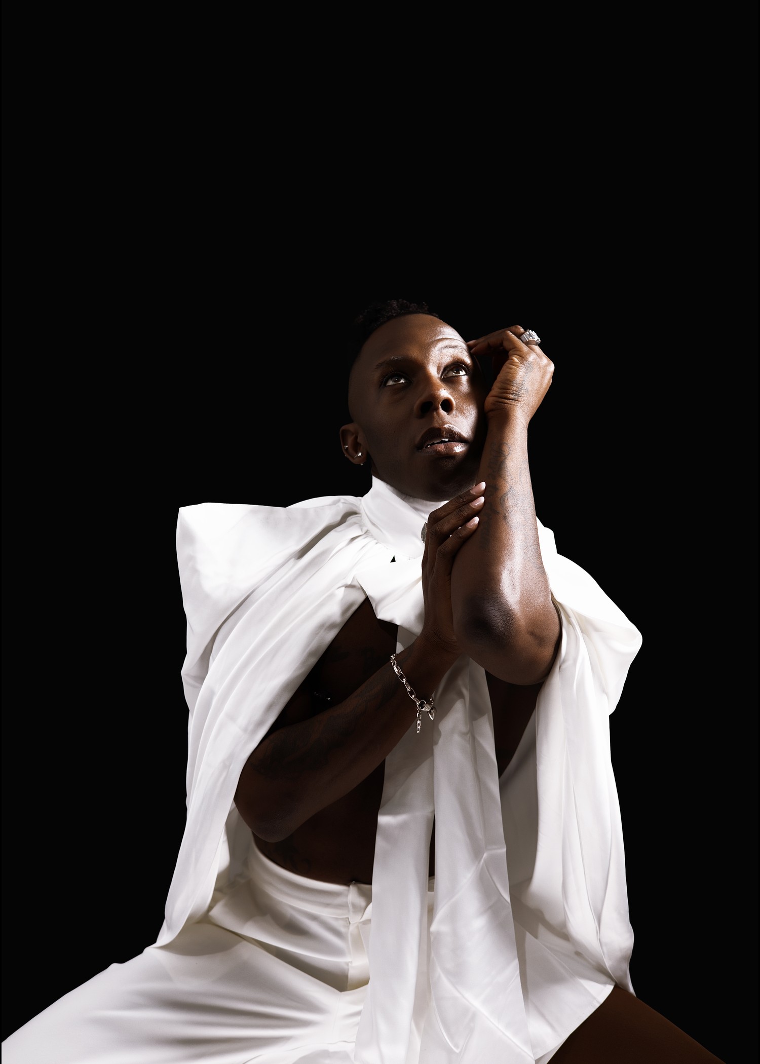 Black male model in white draped garment seated against black background in artistic fashion portrait.