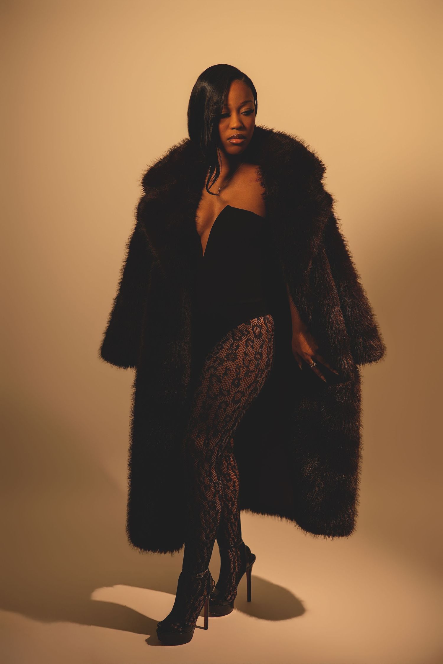 A woman in a long black fur coat, black bodysuit, and leopard tights poses in warm studio light.