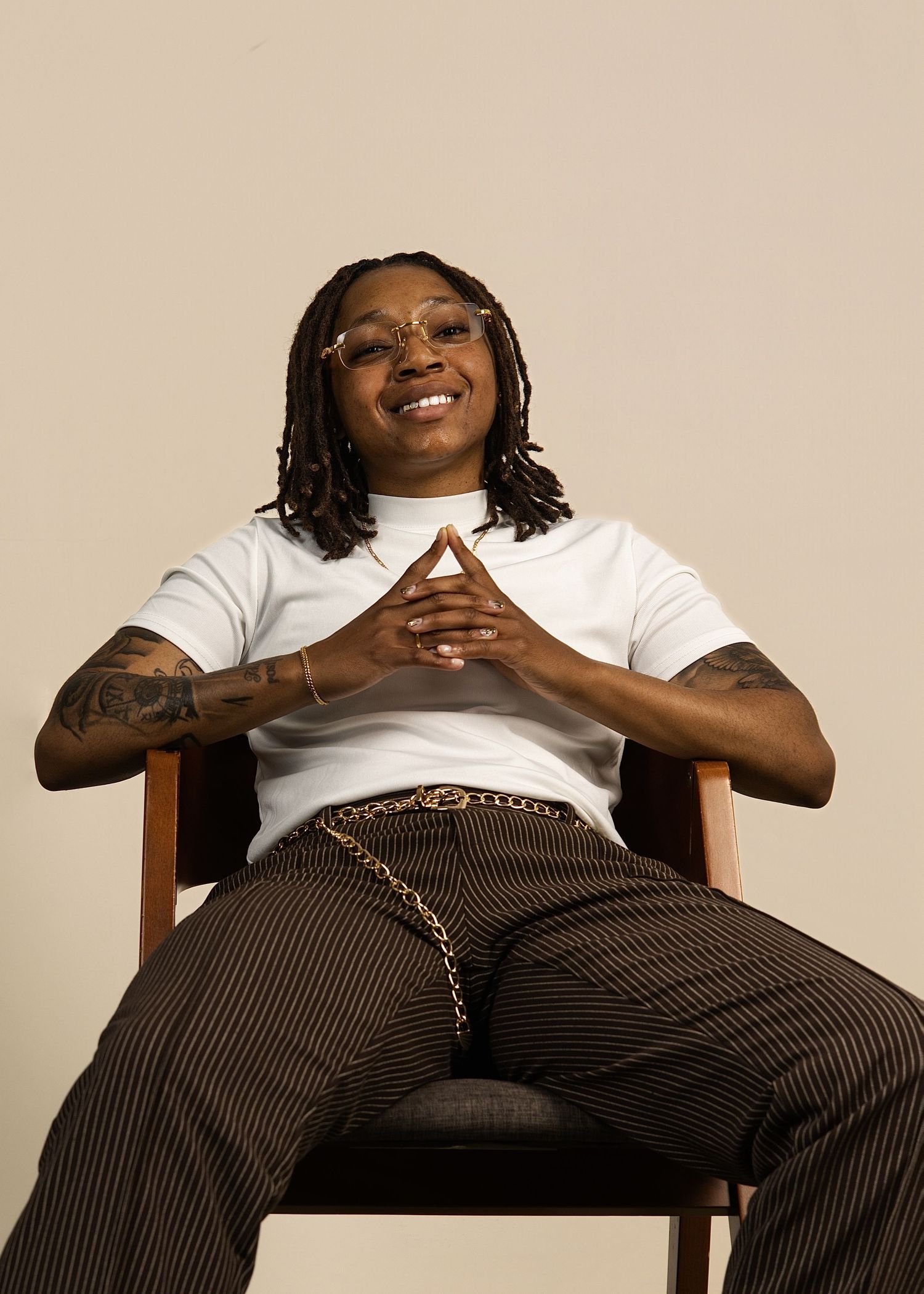 Smiling person with locs and tattoos sits relaxed in wooden chair, neutral background.