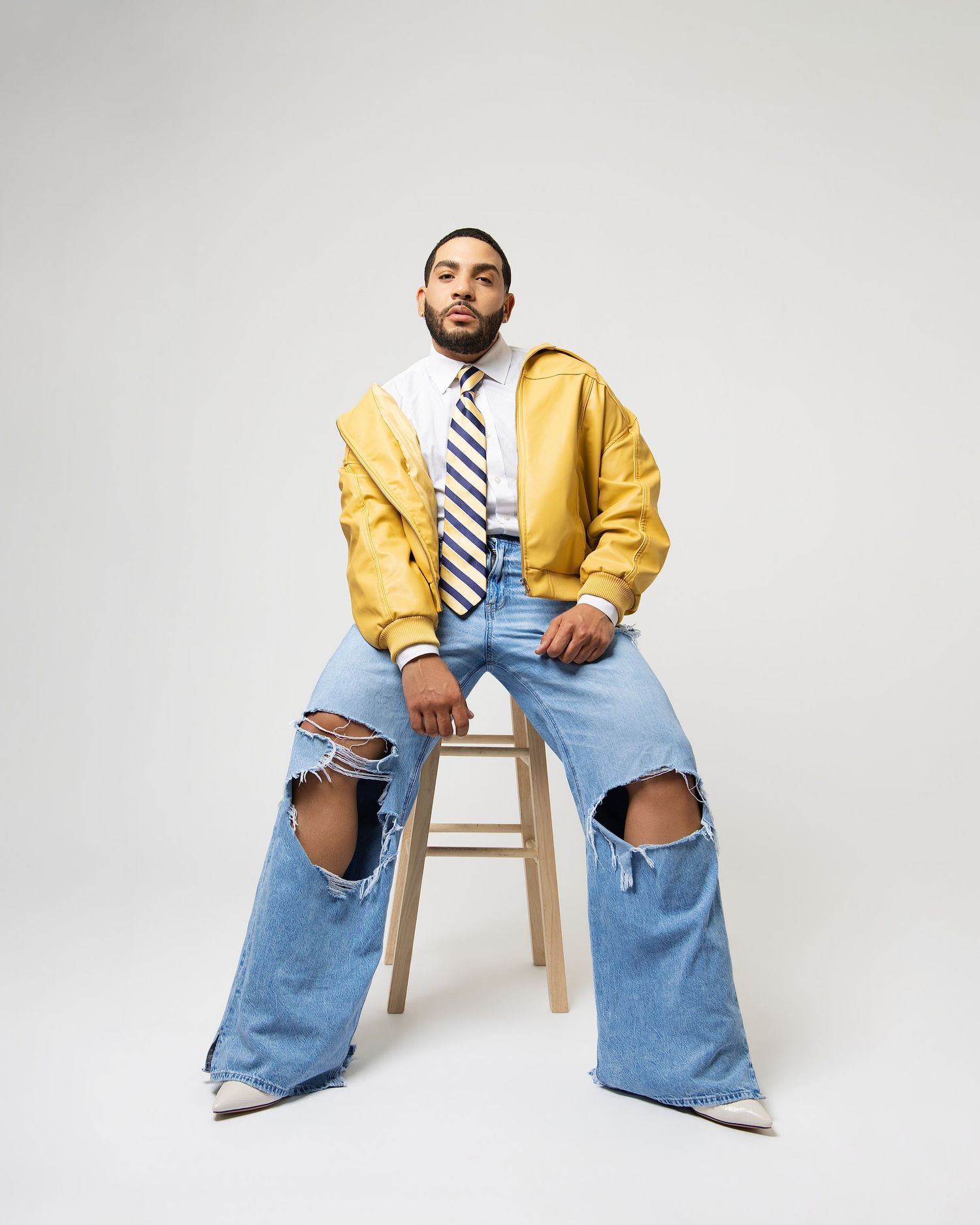 Full body shot of man in yellow jacket, striped tie, and ripped wide-leg jeans seated on wooden stool.