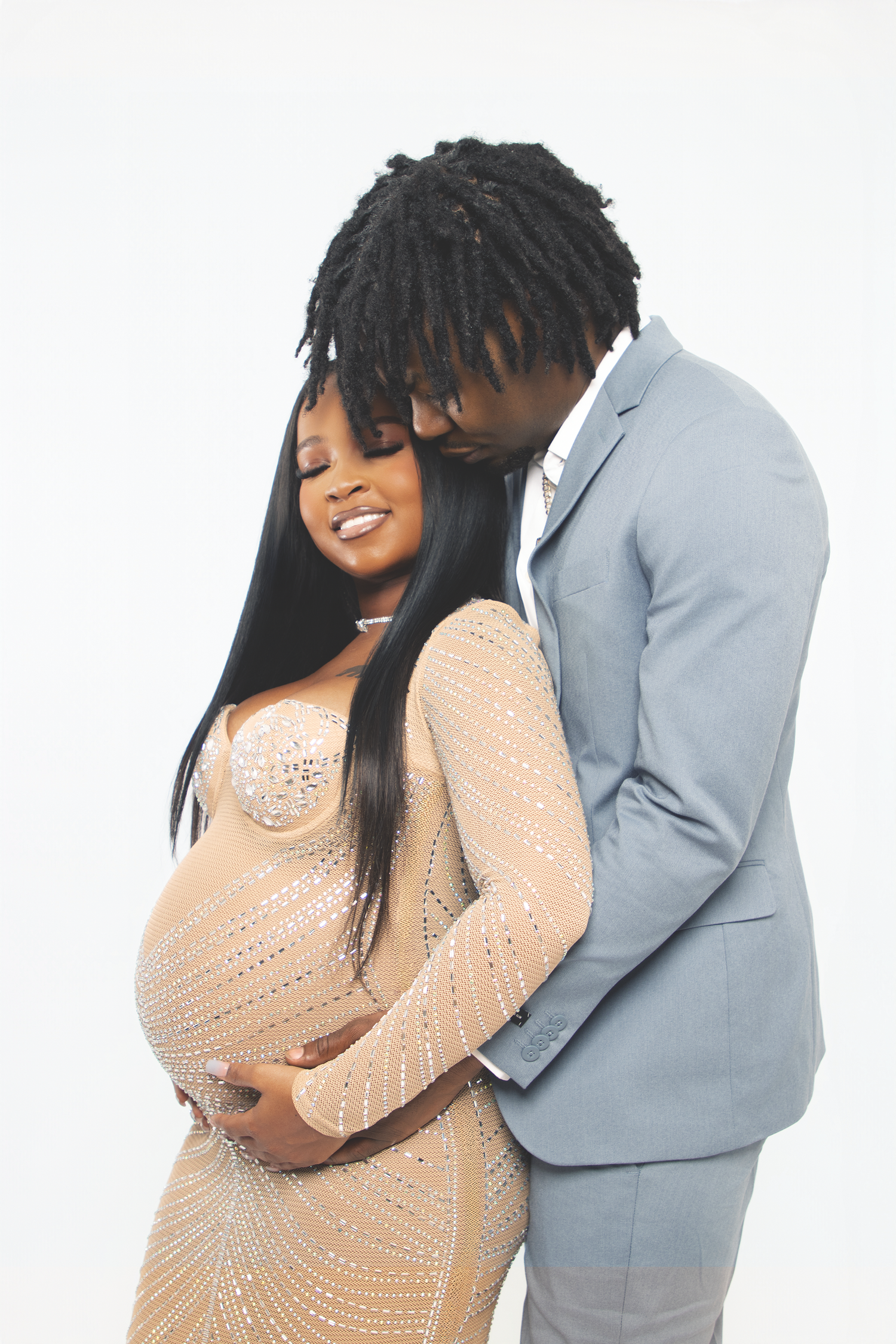 Couple in maternity photoshoot, man in gray suit embracing pregnant woman in beige gown.