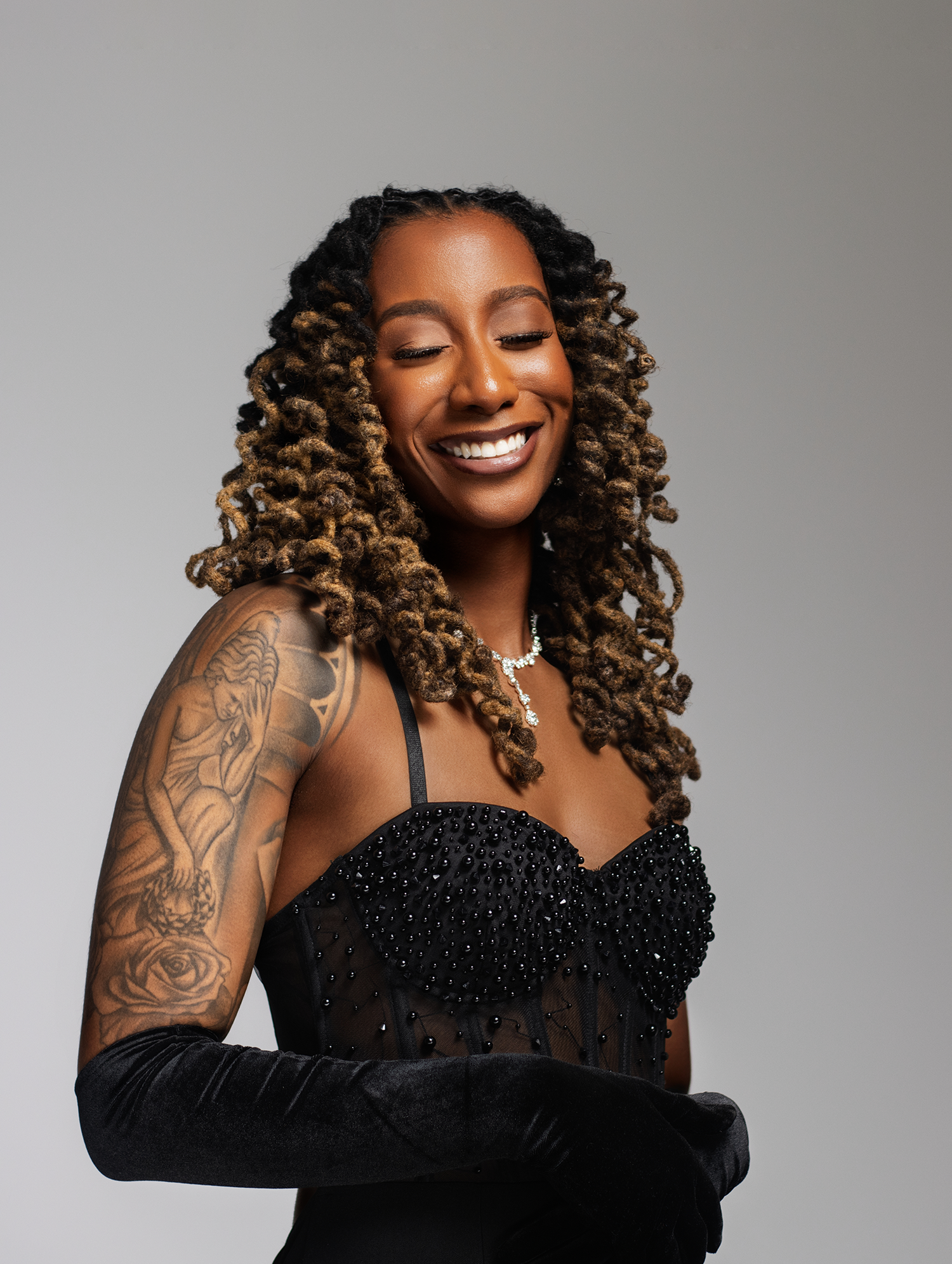 Woman in black beaded bustier gown with long gloves and tattooed arm smiling against gray background.