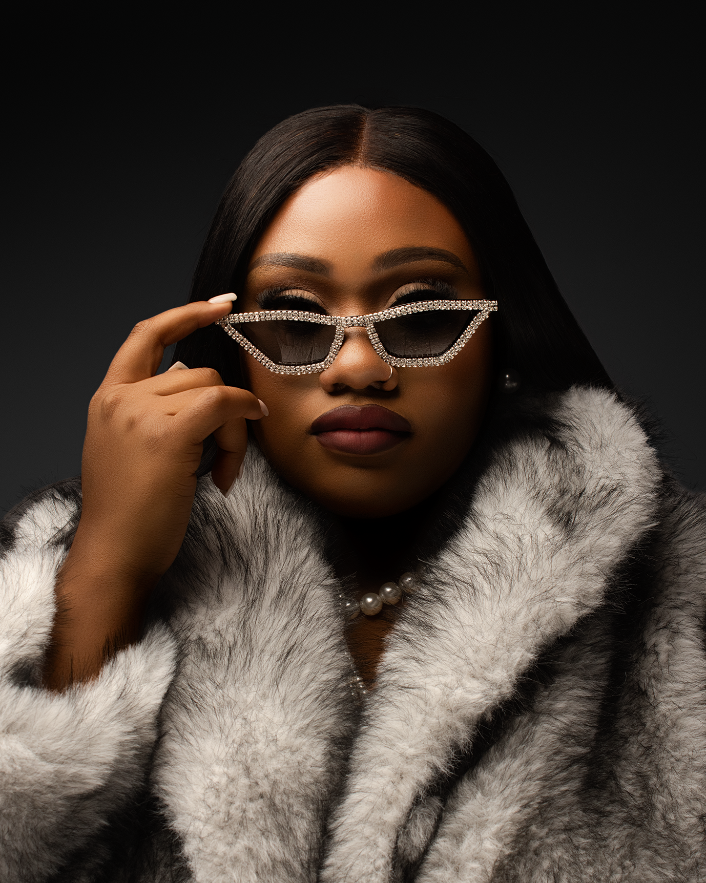 Woman in gray fur coat adjusts rhinestone cat-eye sunglasses in dramatic dark studio portrait.