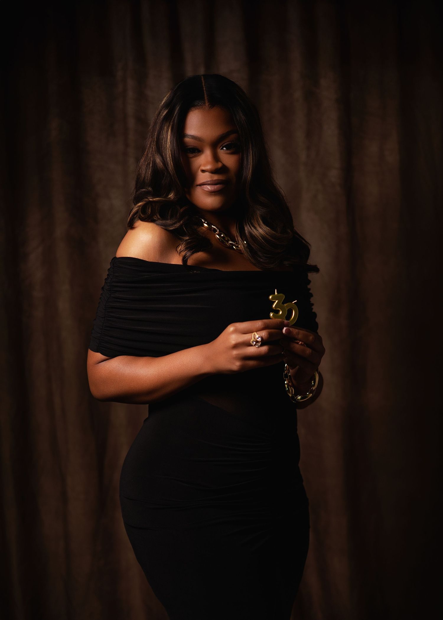 Black woman in black off-shoulder ruched dress holding gold jewelry in classic portrait studio.
