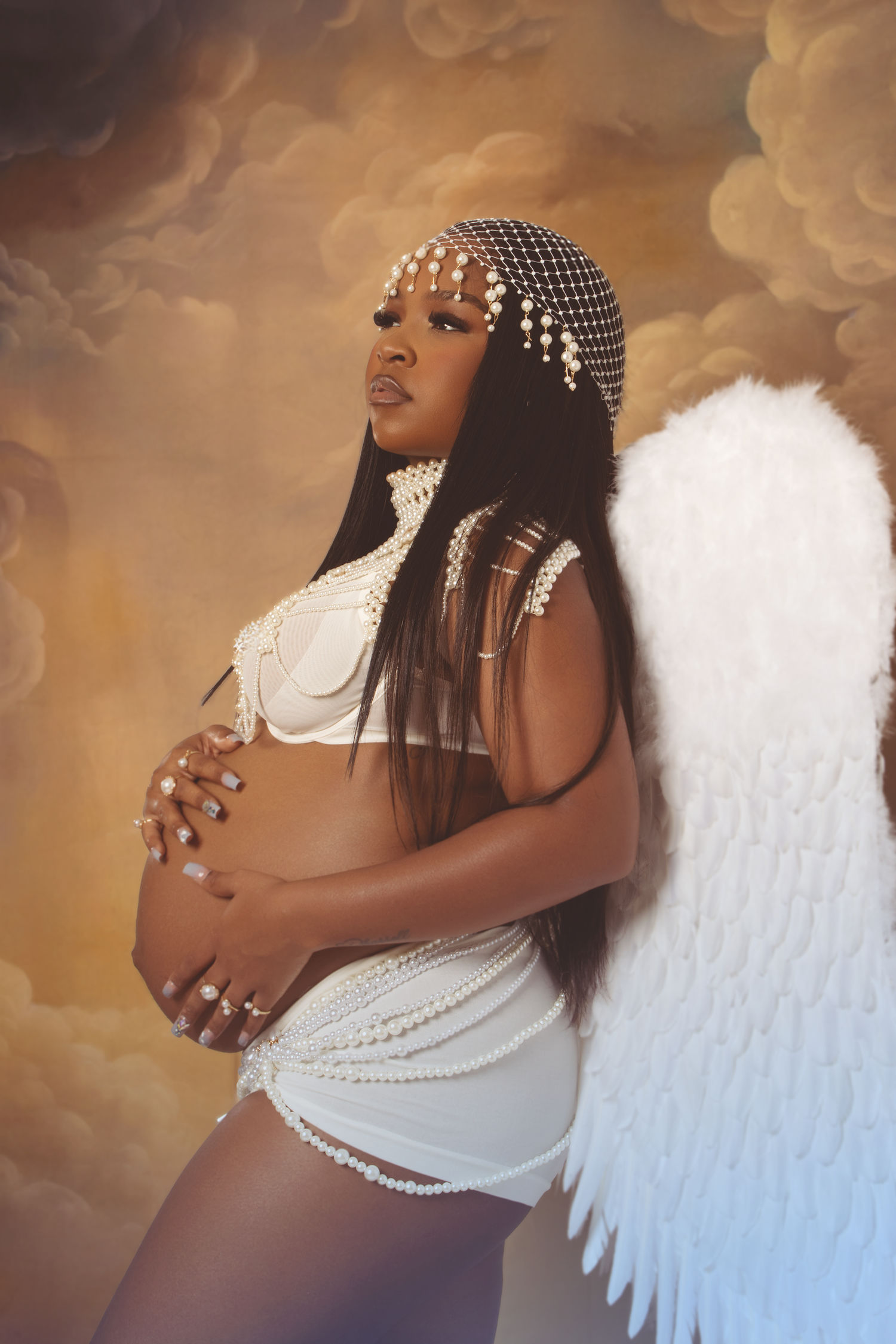 Pregnant woman poses as an angel with white wings and pearl headpiece on cloudy backdrop.