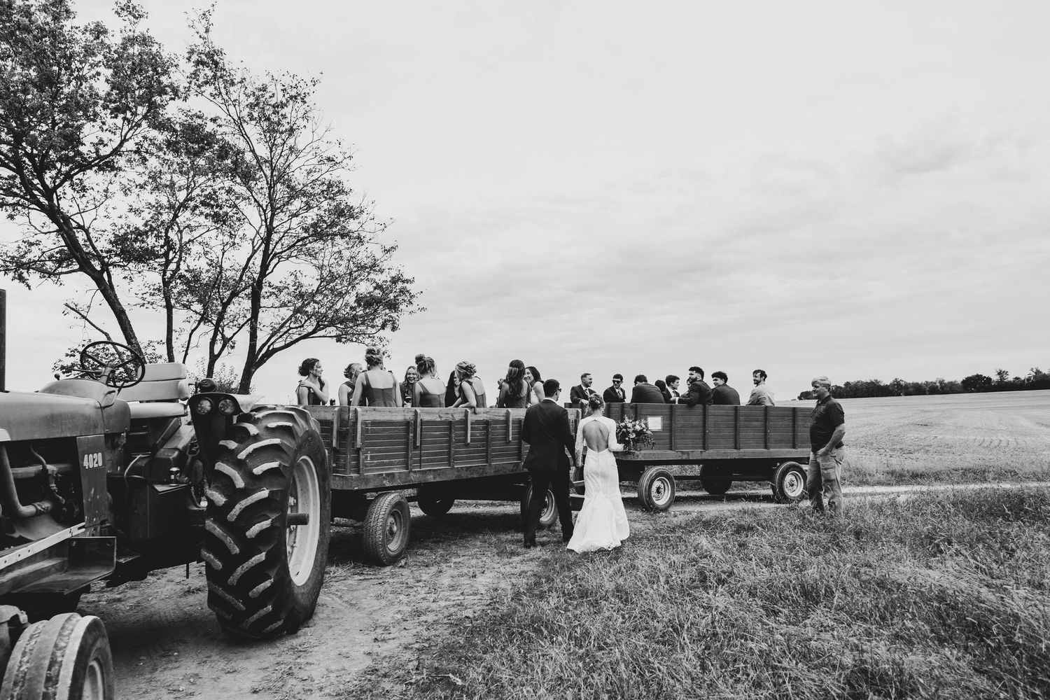 Black and white photo of wedding guests riding on a tractor-pulled wagon through a rural farm field.