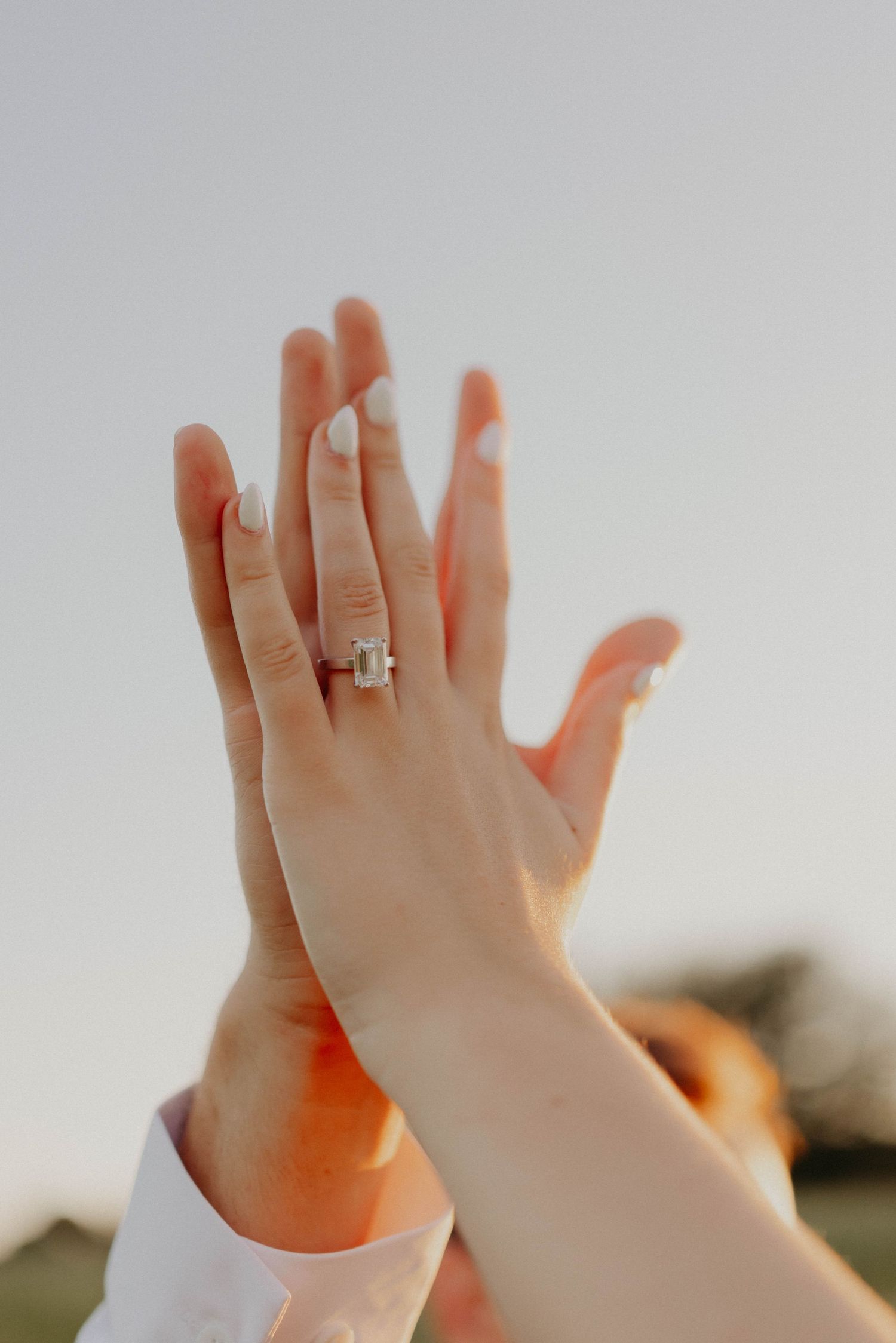 Bride showing off her elegant emerald-cut diamond wedding ring while holding hands outdoors at sunset.